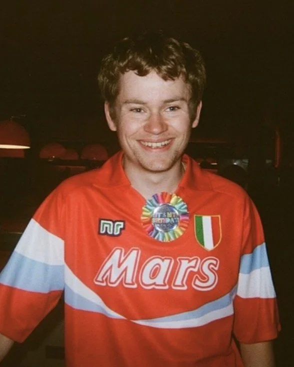 A young man smiling, wearing a red sports jersey with white and light blue stripes, with a Laurel wreath medal, and patches including an Italian flag and the word 'Mars'.