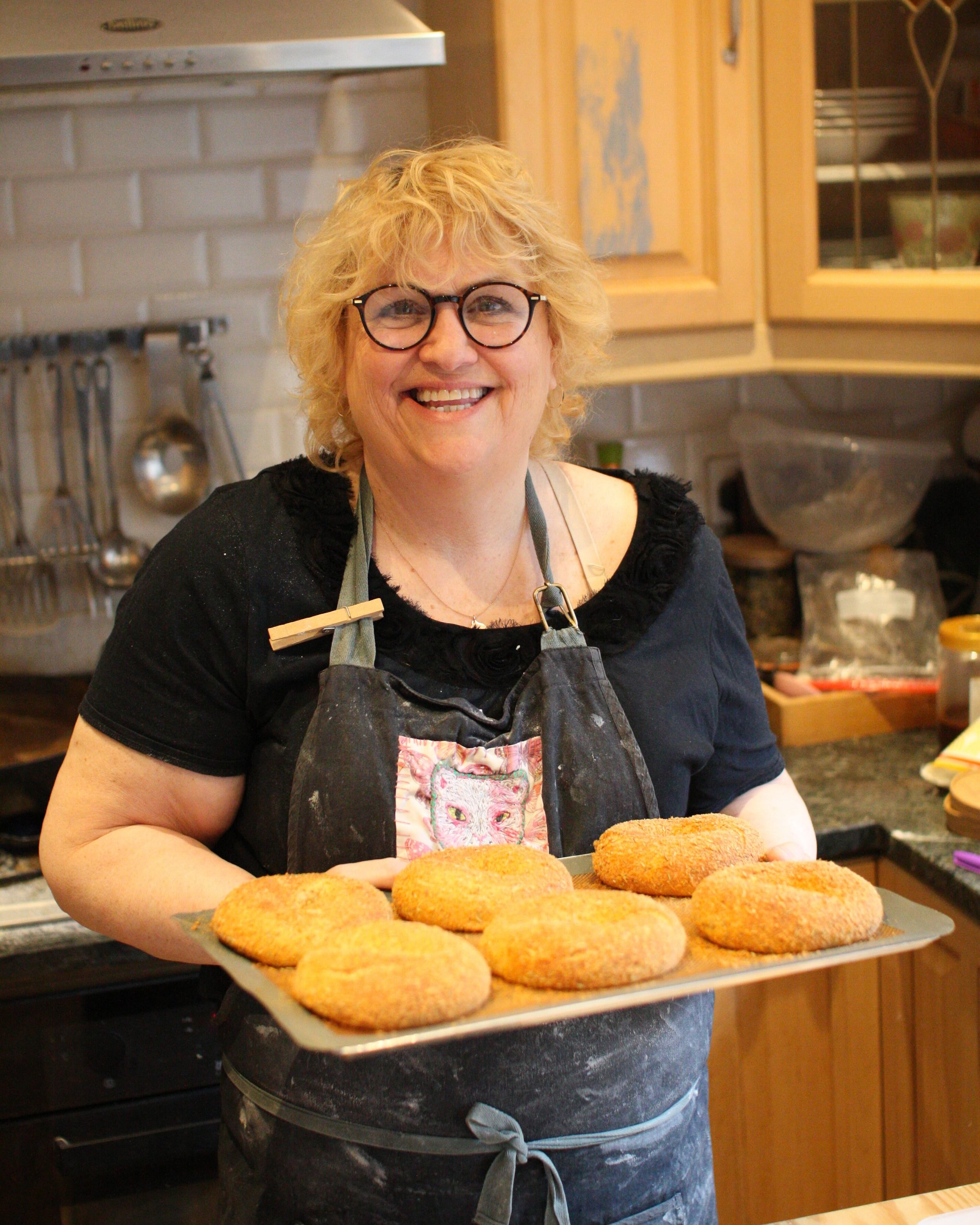 A woman with curly blonde hair and glasses standing in a kitchen, holding a tray of baked breaded patties.