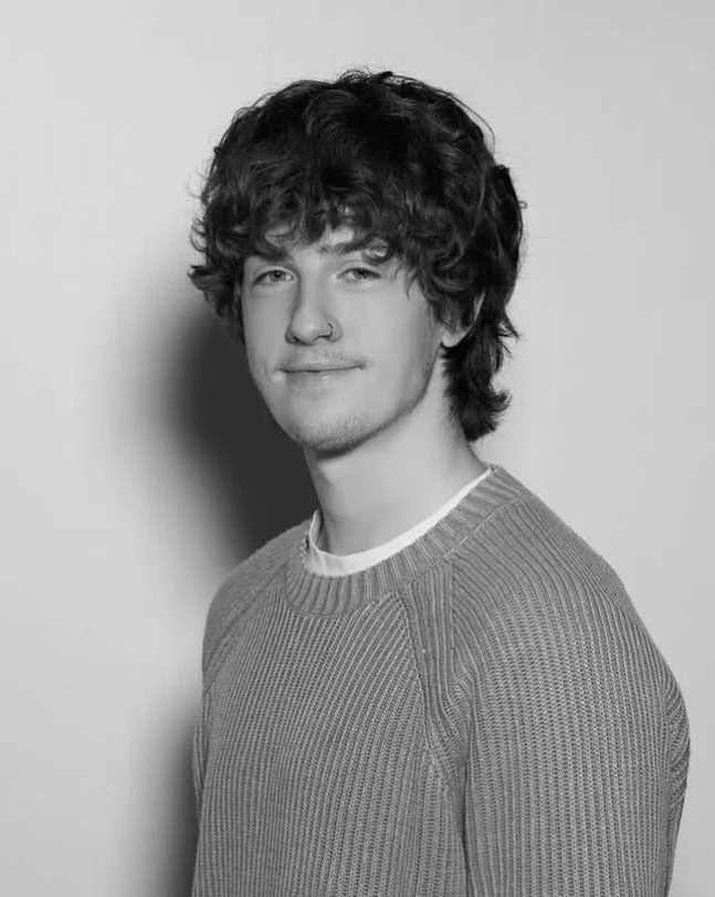 Black and white portrait of a young man with curly hair, wearing a ribbed sweater, looking at the camera with a slight smile.