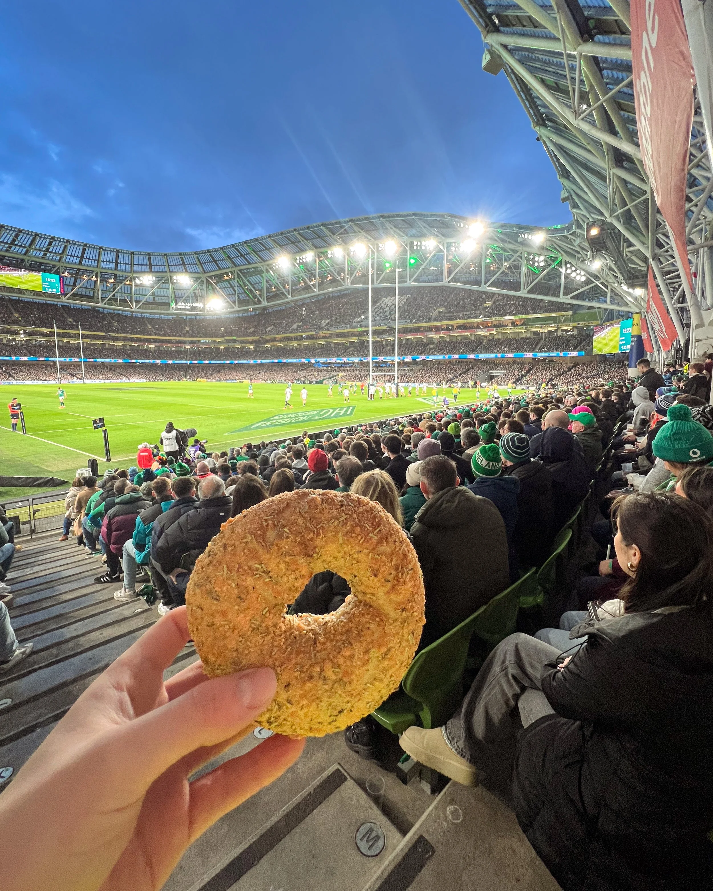 A person holding a bagel in the foreground at a rugby stadium filled with spectators, with players on the field under bright lights and a clear evening sky.