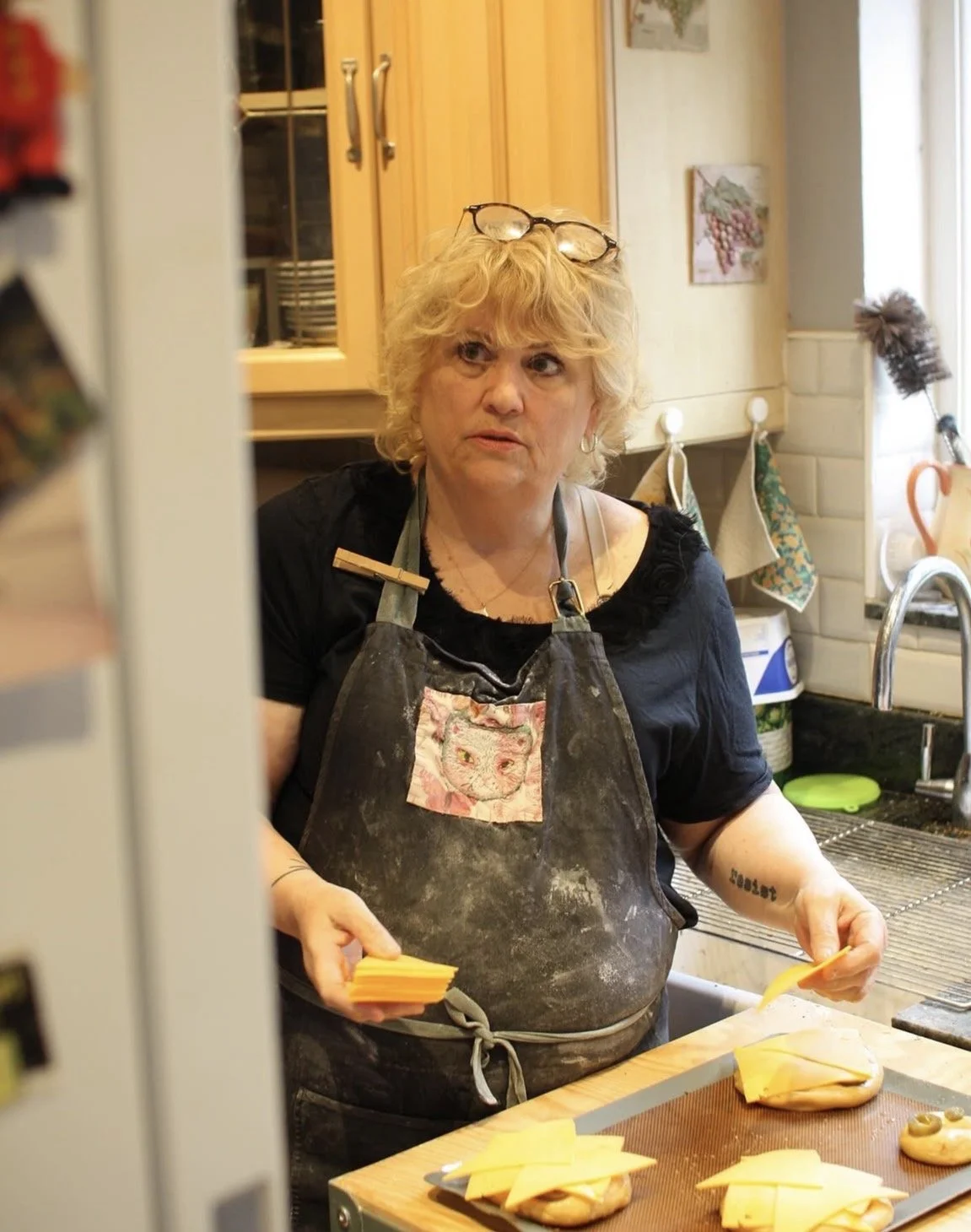 A woman with glasses on her head, wearing a dark shirt and an apron, preparing food in a kitchen. She is holding slices of cheese on a cutting board with a concerned expression, with some bread and cheese on trays in front of her.