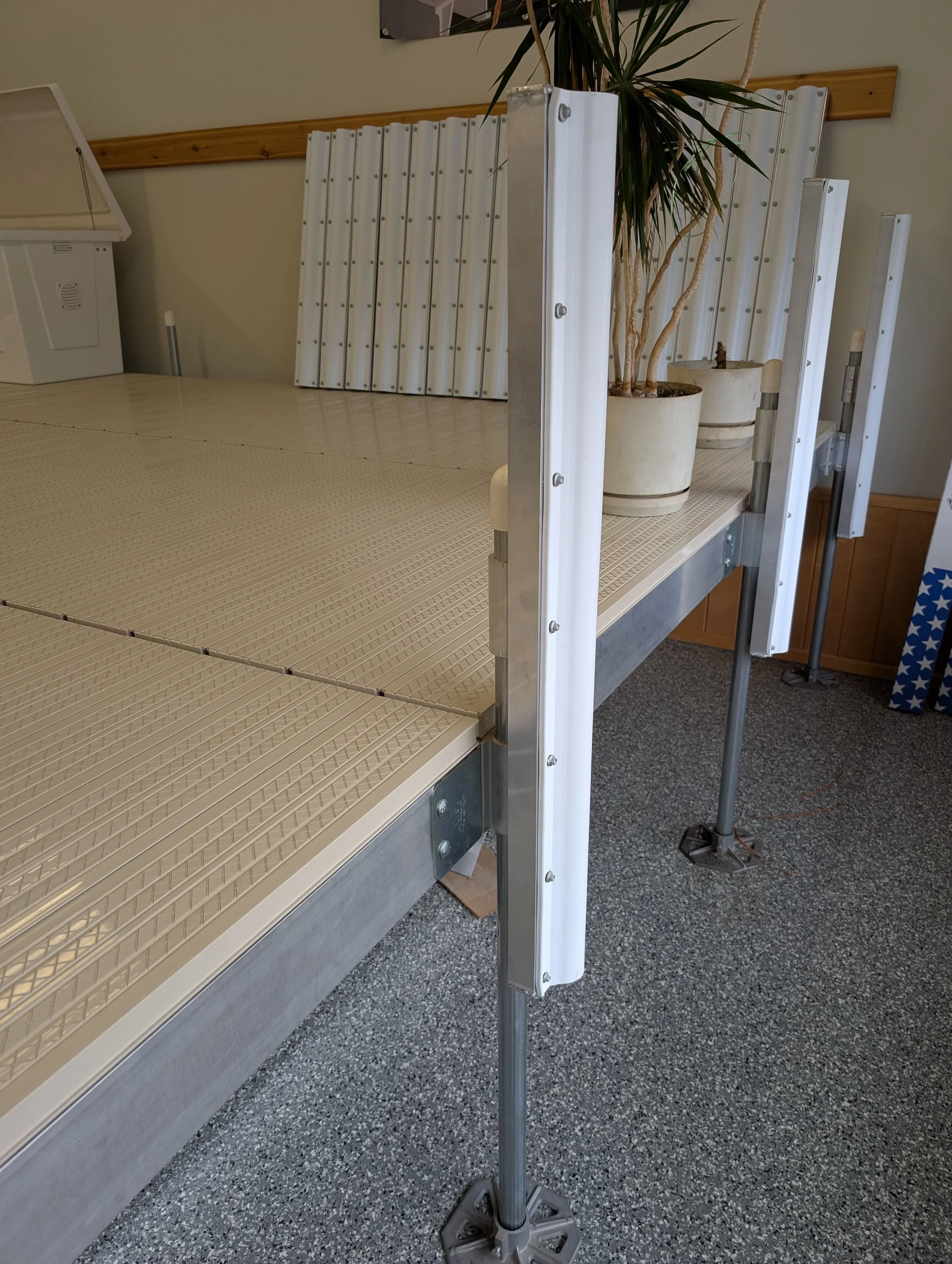 A beige, textured platform with metal edges, supported by a metal frame with vertical supports and casters, set up in an indoor area with a grey speckled carpet floor. Behind the platform, there are metal panels and a tall potted plant with long, narrow leaves.