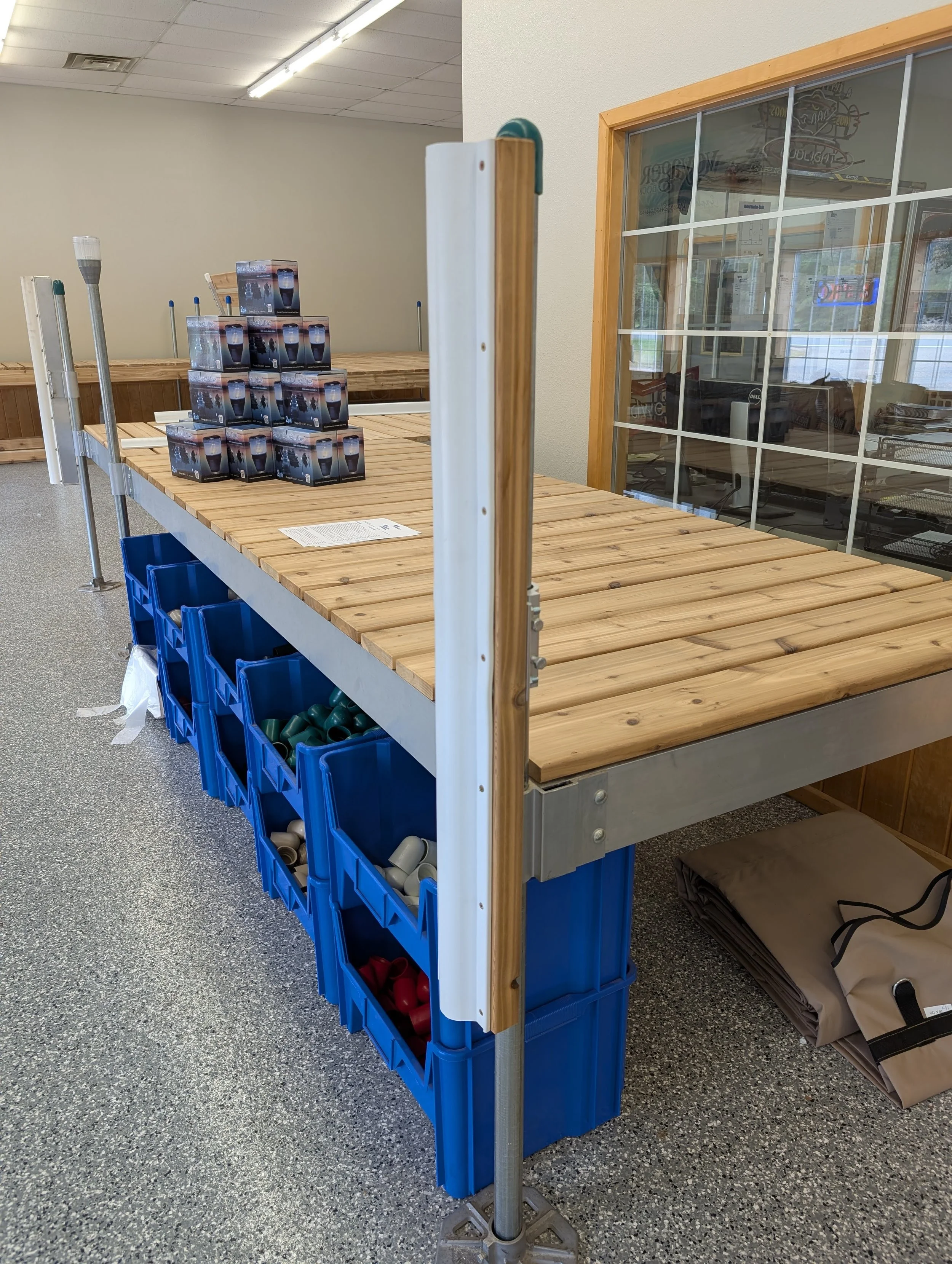 Empty wooden checkout counter with blue plastic bins underneath, some containing shoes and other items, in a retail store with a glass window separated by a wooden frame, and a paper on the counter.