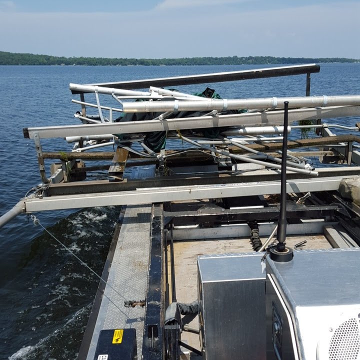 Close-up of a boat or pontoon on the water with metal pipes and equipment, and a body of water in the background.
