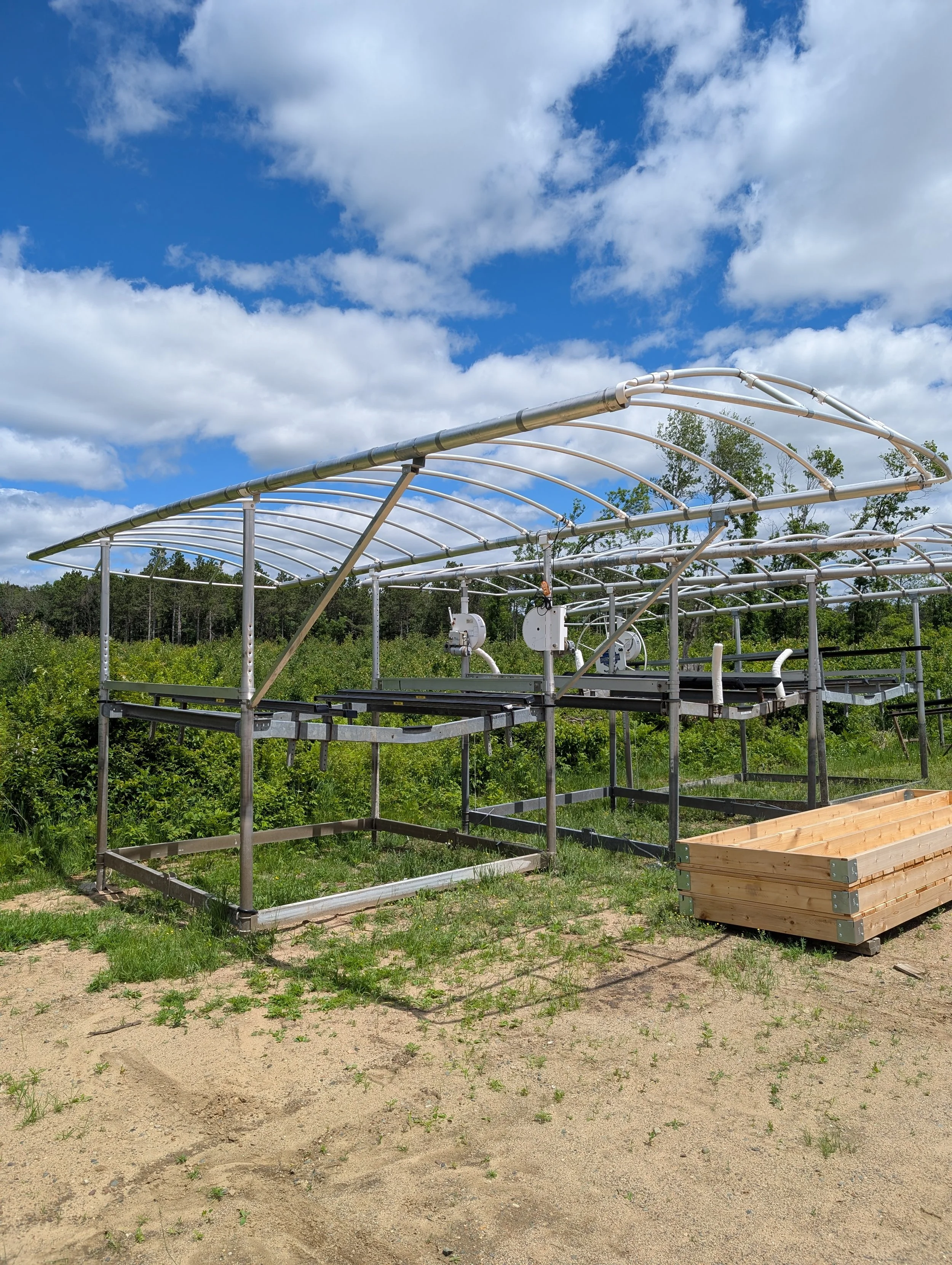 An outdoor greenhouse structure with a metal frame and pipes, set in a green field with trees and a bright blue sky with scattered clouds overhead.