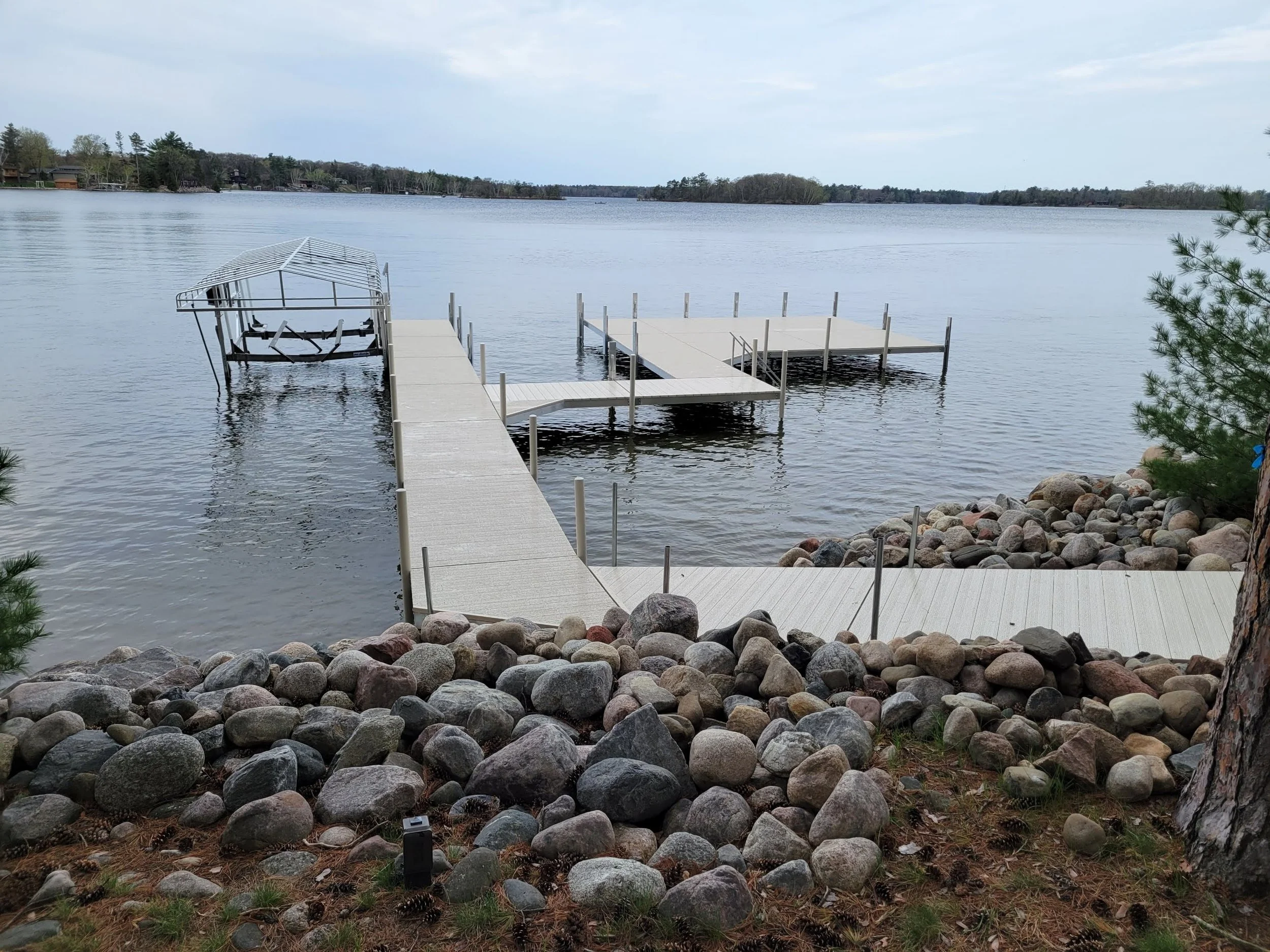 A lakeside scene with a long dock extending into the water, a boat lift with a boat cover on the left, and a rocky shoreline with a small tree in the foreground. There are houses and trees across the lake in the background under a cloudy sky.