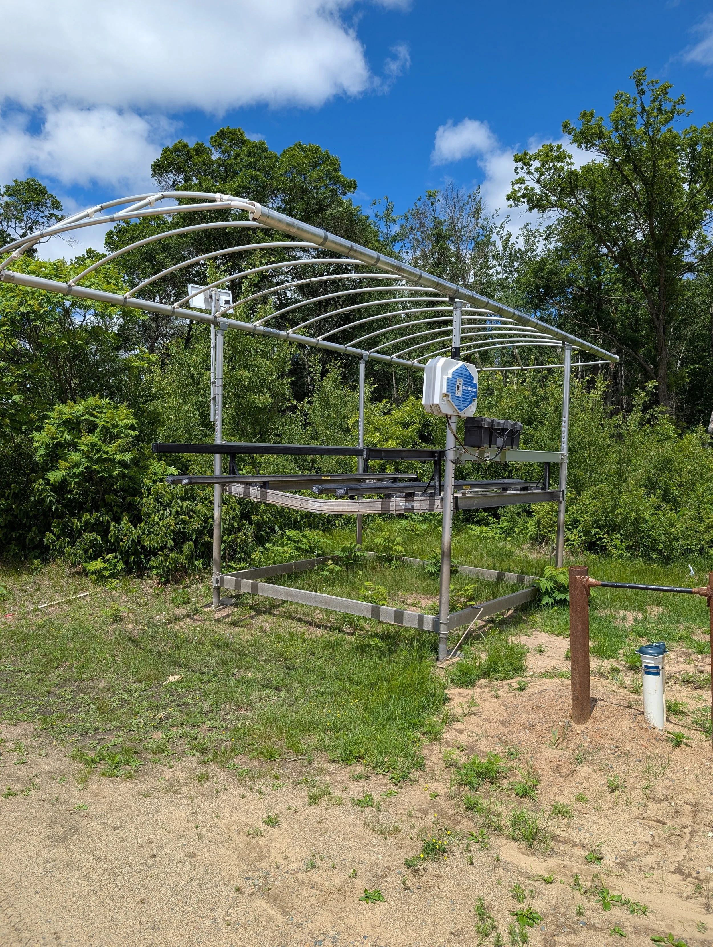 A partially constructed greenhouse frame made of metal pipes is set up on grassy soil with trees in the background. Sunlight and scattered clouds are visible in the blue sky.