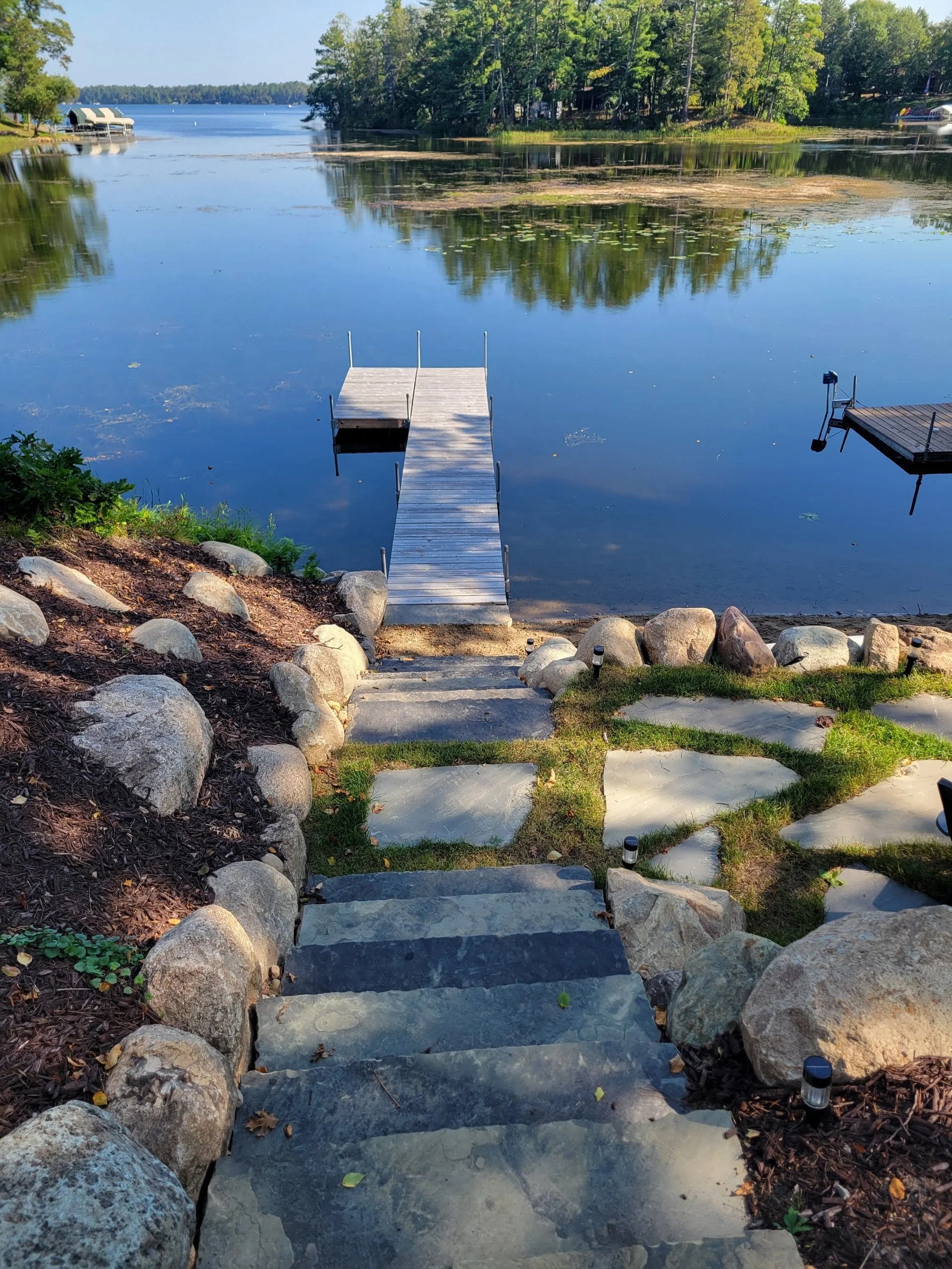Steps leading down to a dock on a peaceful lake, surrounded by rocks and greenery, with trees and boats visible in the background.