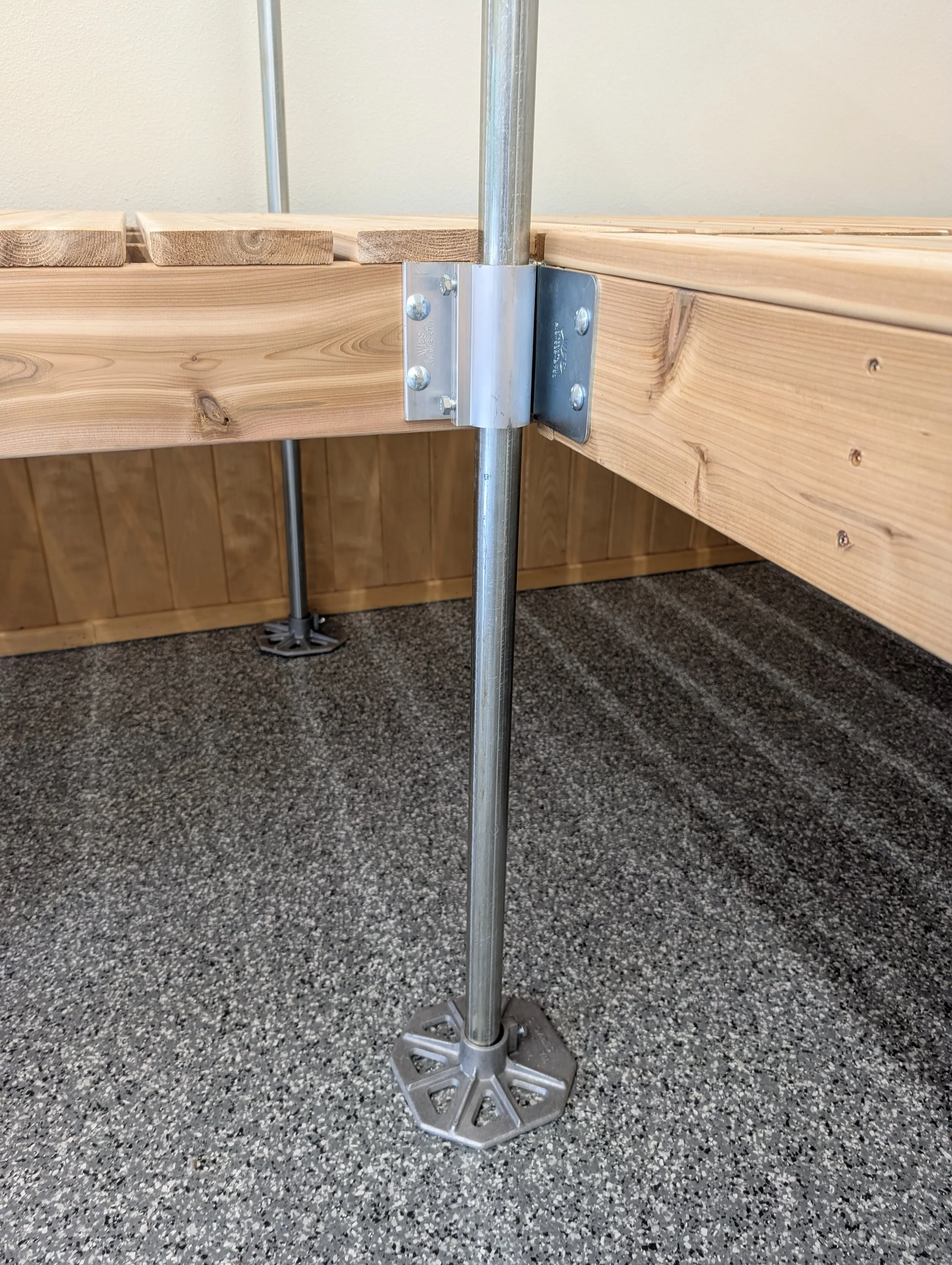 Close-up of a metal scaffold support pole with a clamped metal plate, attached to a wooden platform with a wooden edge and a beige wall in the background. The flooring is speckled gray.