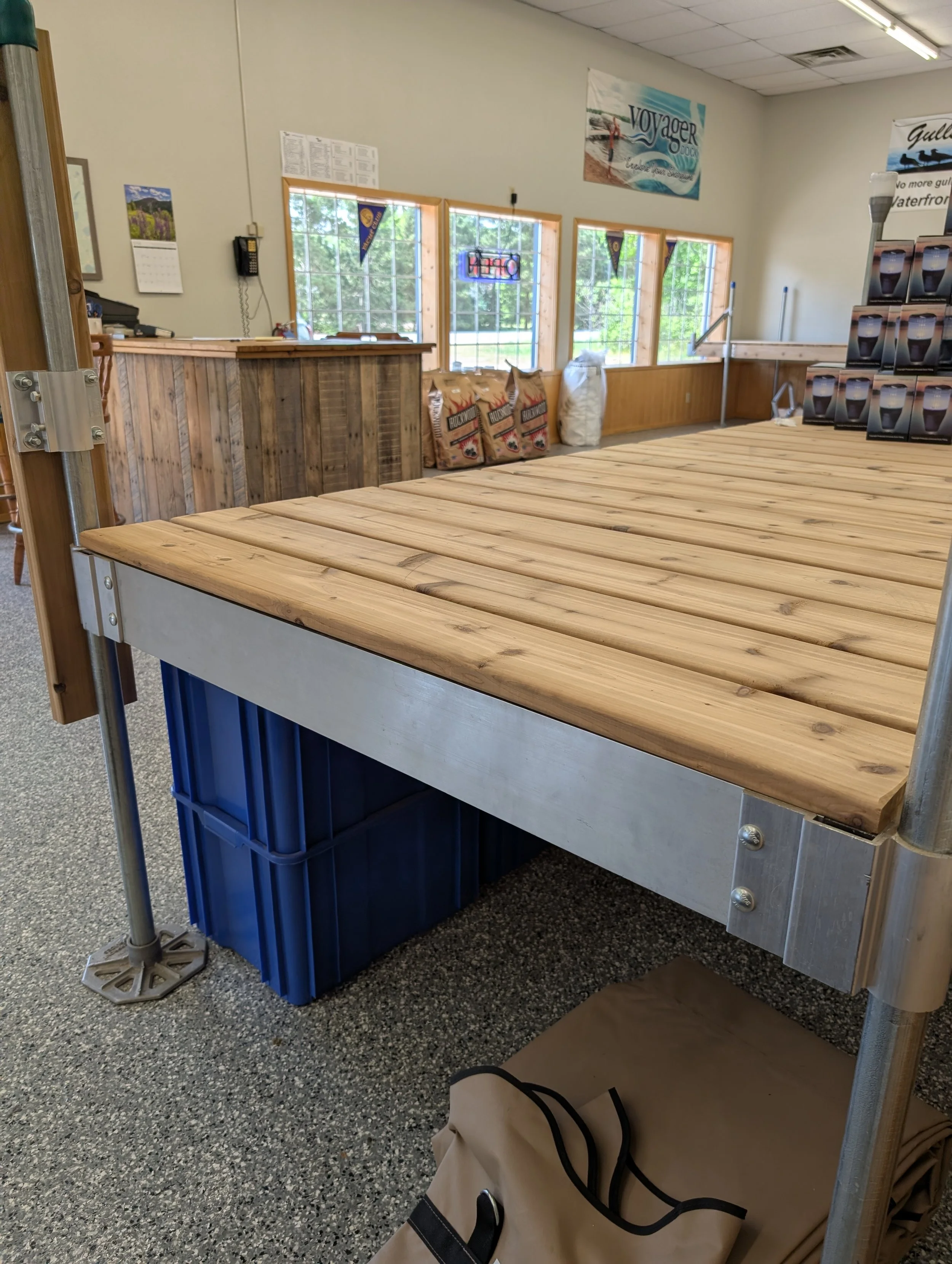 Image of an indoor space with a wooden table, a blue plastic crate, a paper bag, and multiple bags of charcoal near a wooden bar counter with three windows behind it, an open sign, and promotional banners on the wall.