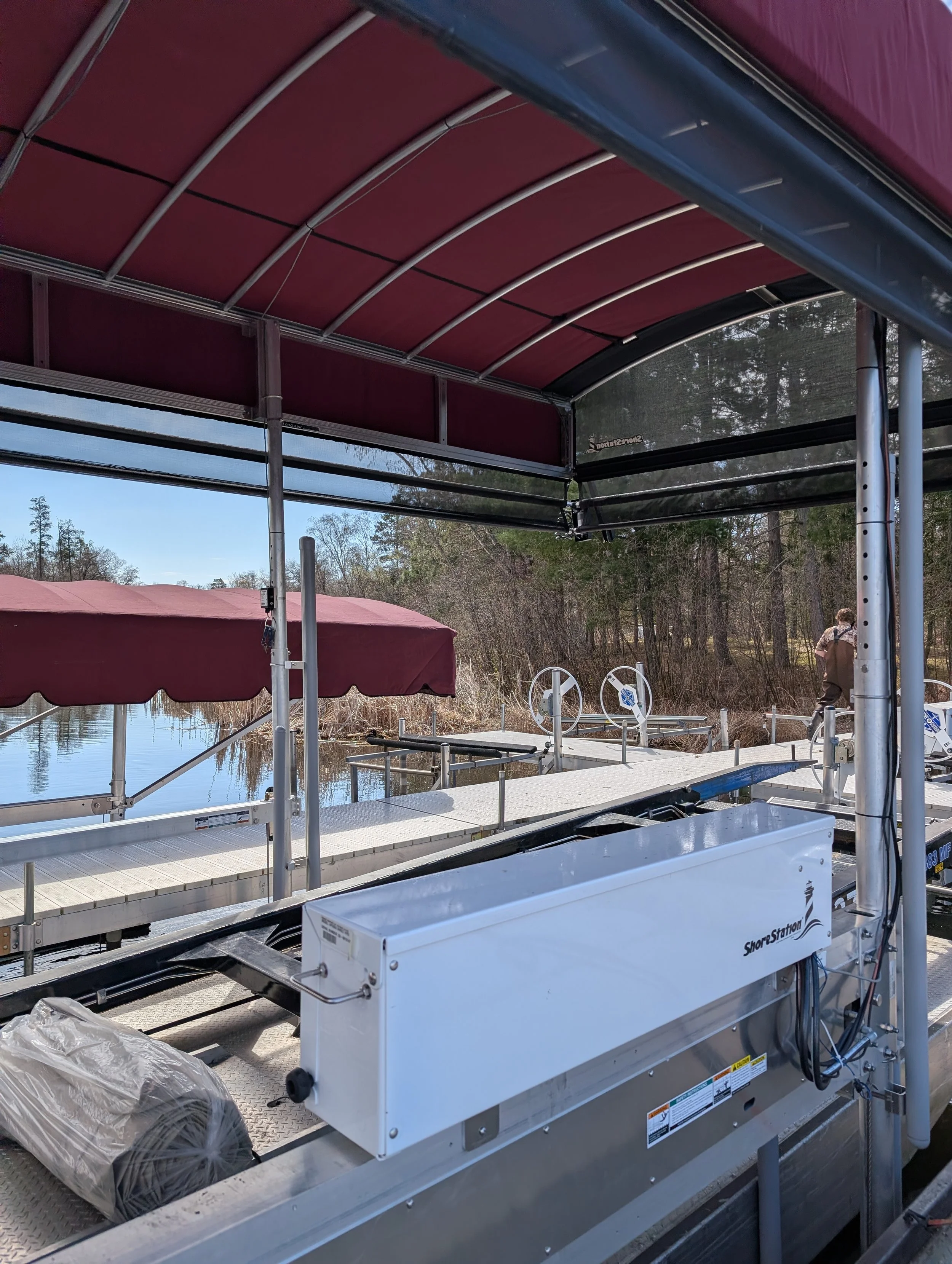A boat dock with a metal, red canopy, and a white ShoreStation boat lift. The lift is empty except for some plastic wrapping. In the background, a man is standing near the water with trees and a cloudy sky.