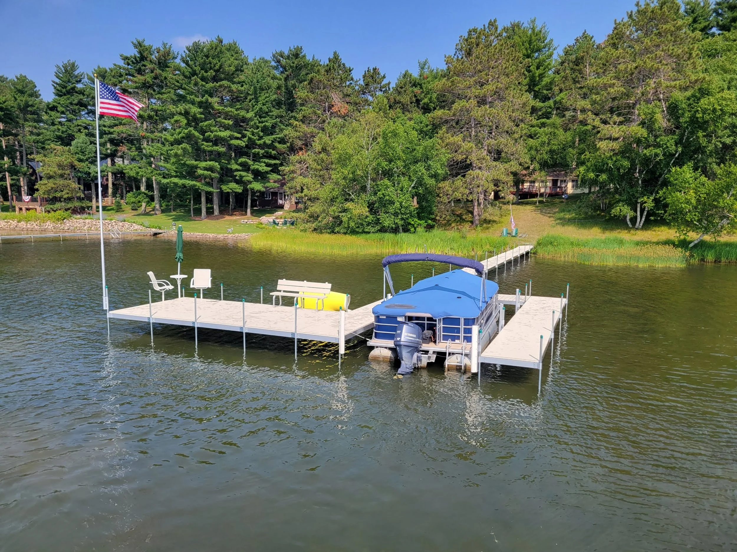 A lakeside scene featuring a dock extending over the water with a covered boat, outdoor chairs, and umbrellas. An American flag flies on a tall pole, and lush green trees and houses are visible in the background under a clear blue sky.