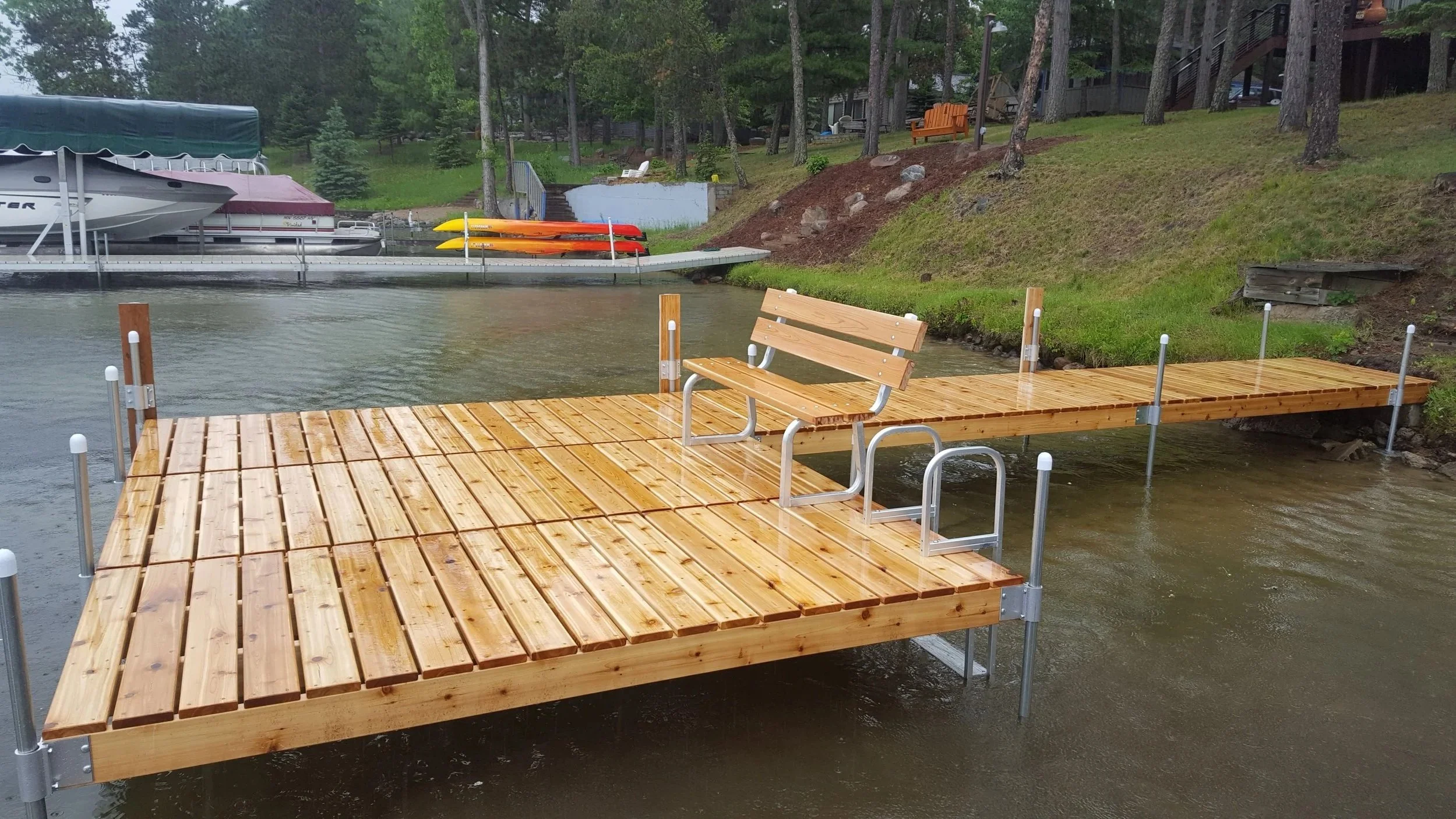 Wooden dock extending into a lake with metal handrails, surrounded by water and greenery, with boats and kayaks stored on a pier
