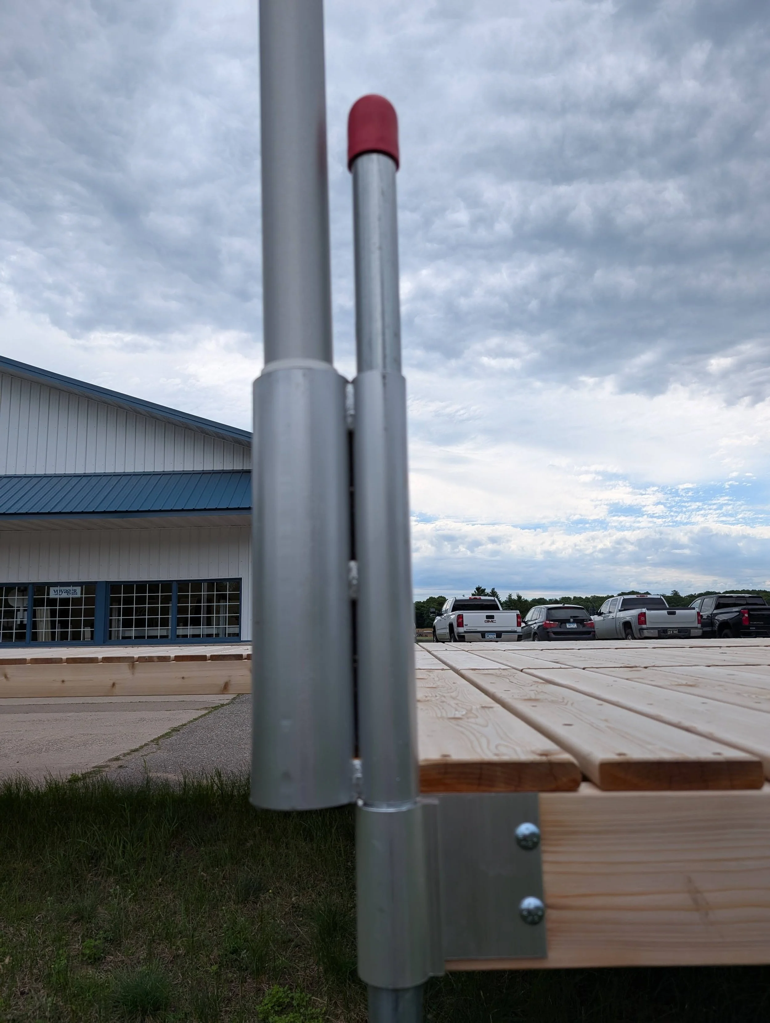 A close-up view of a wooden outdoor picnic table with a metal frame, set against a parking lot with several pickup trucks, and a building with a metal roof and siding. The sky is cloudy.