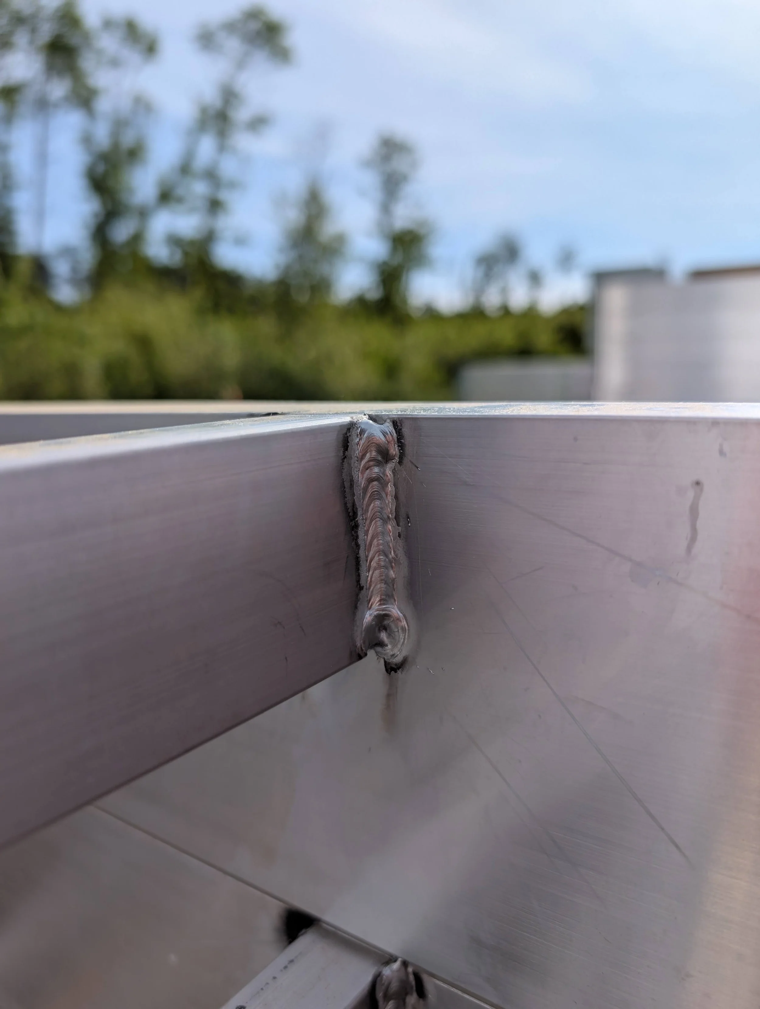 Close-up of a metal corner joint with a weld, showing a slightly bent sheet metal surface against a blurred background of trees and blue sky.