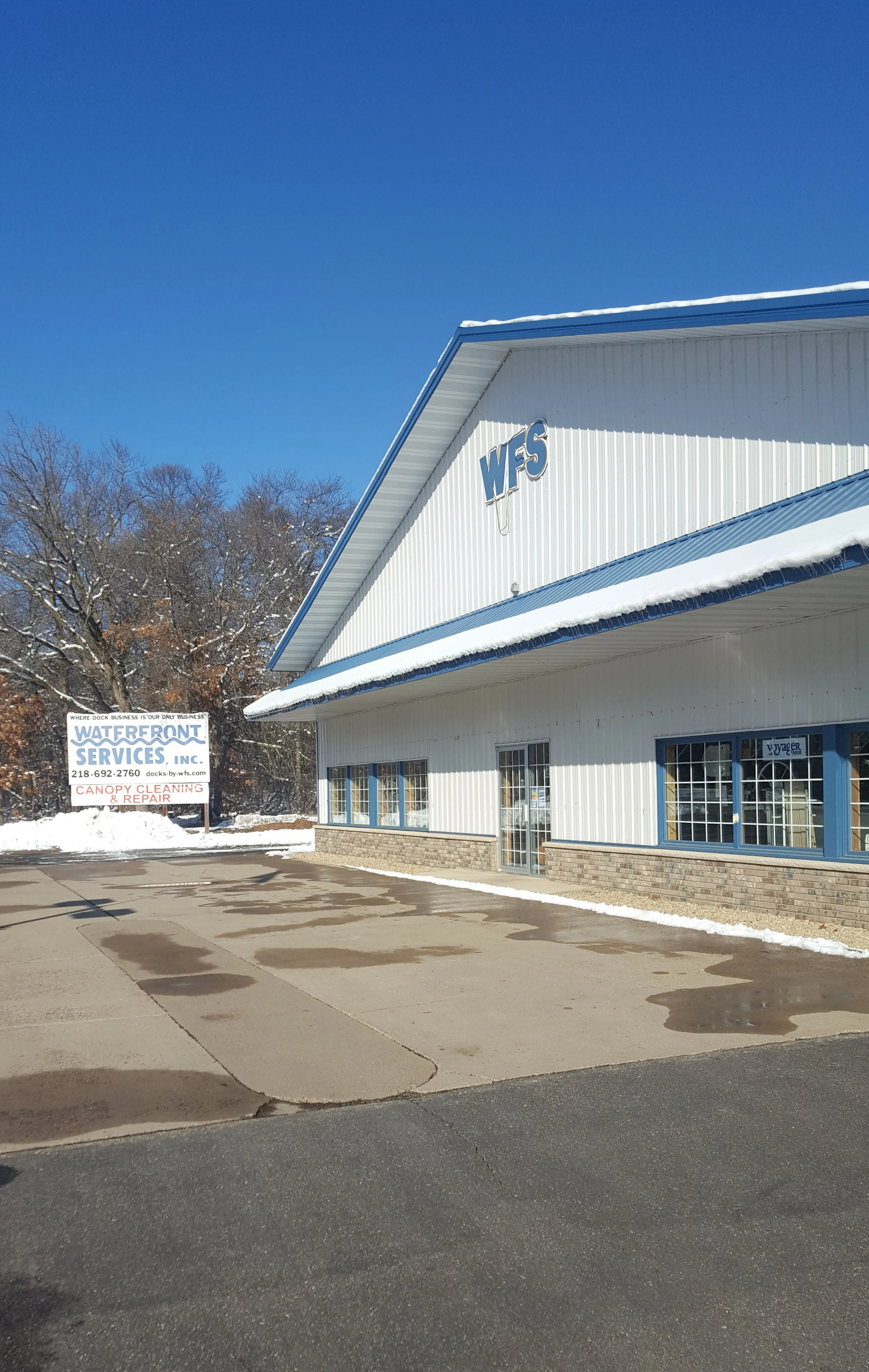White building with blue accents and the letters "WFS" on the facade, snow on roof, blue sky, with a sign for Waterfront Services, Inc., offering dock business and canopy cleaning and repair, in front of some trees with winter foliage.
