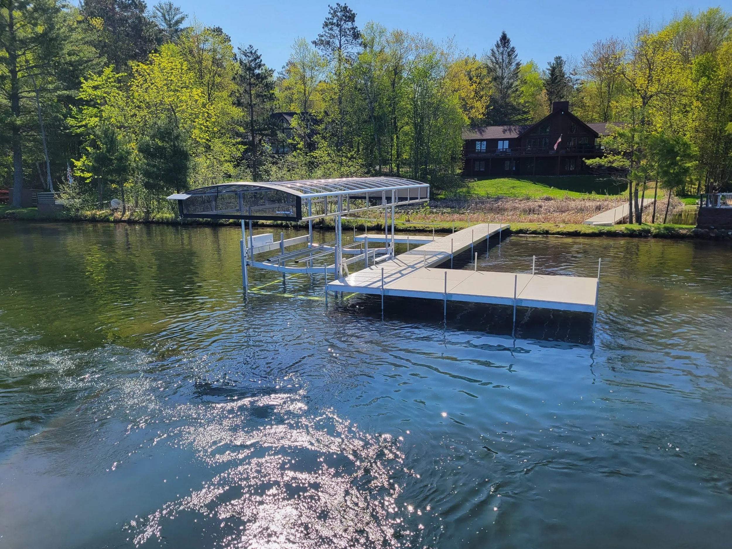 A lakeside scene featuring a dock and boat lift in the foreground, surrounded by lush green trees and a dark wood house on a hill in the background on a sunny day.
