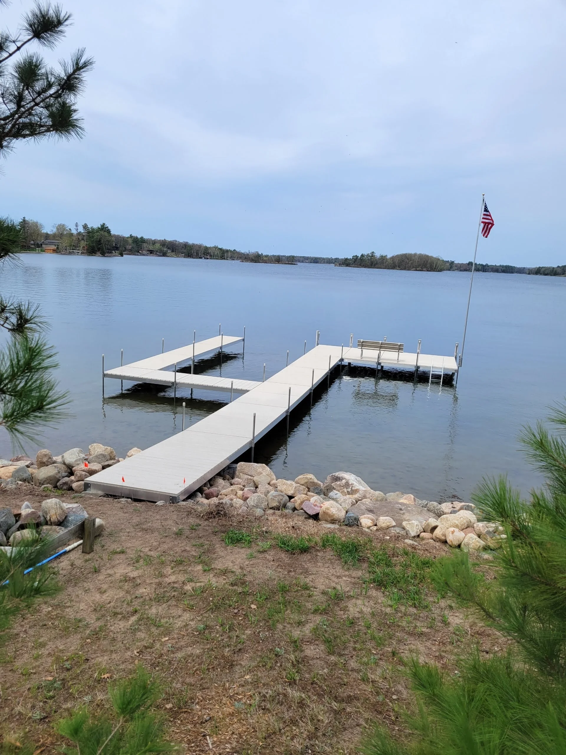 A lakeside scene with a floating dock and American flag on a pole, surrounded by trees and rocks, under a cloudy sky.
