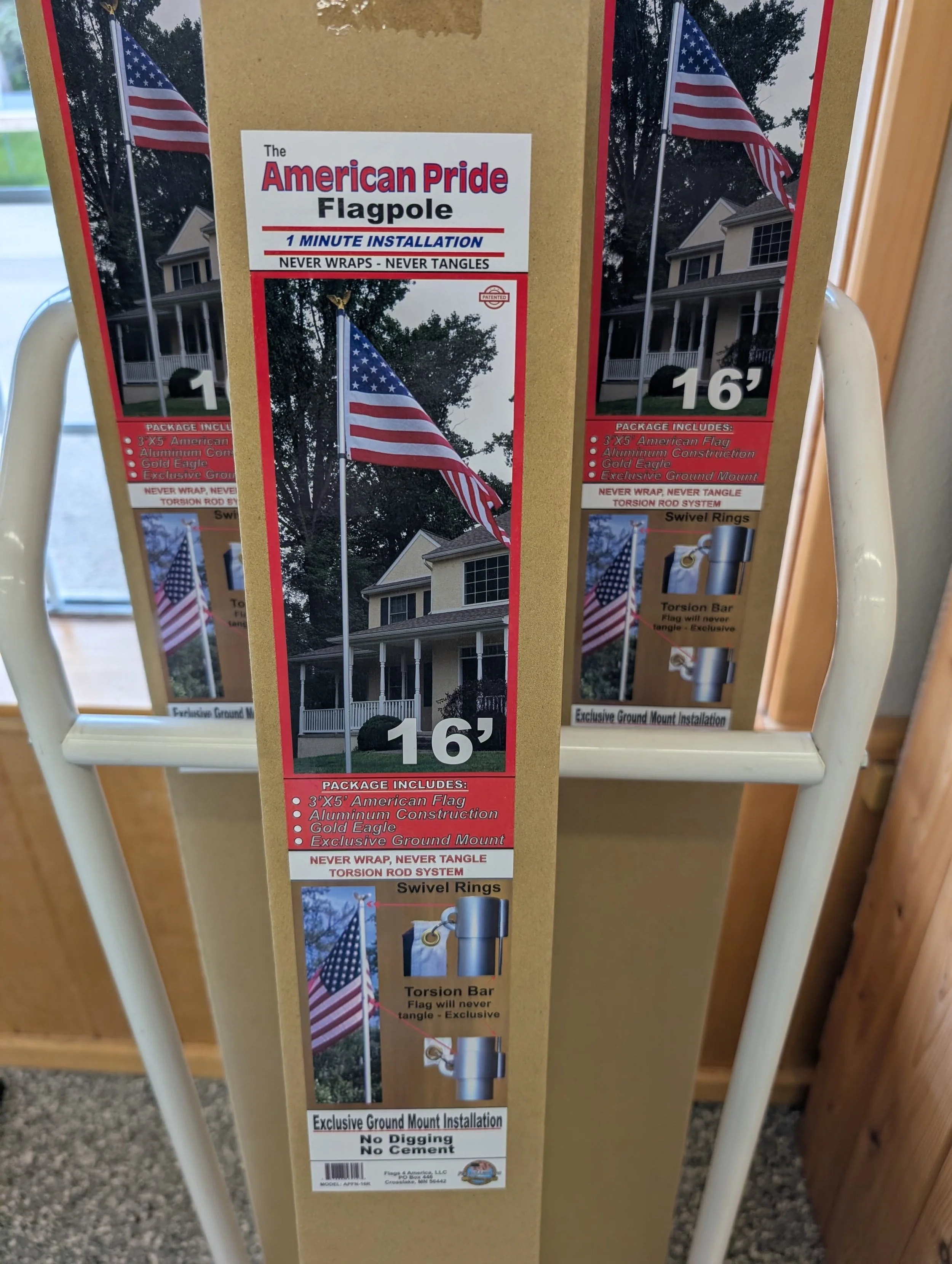 Box for an American Pride flag featuring the flag waving on a pole in front of a house with trees in the background. The packaging mentions a 1 minute installation, no wrapping or tanging, and an exclusive ground mount system. The box indicates the flag size is 3 by 5 feet, made of aluminum with a gold eagle, and comes with swivel rings and a torsion rod system.