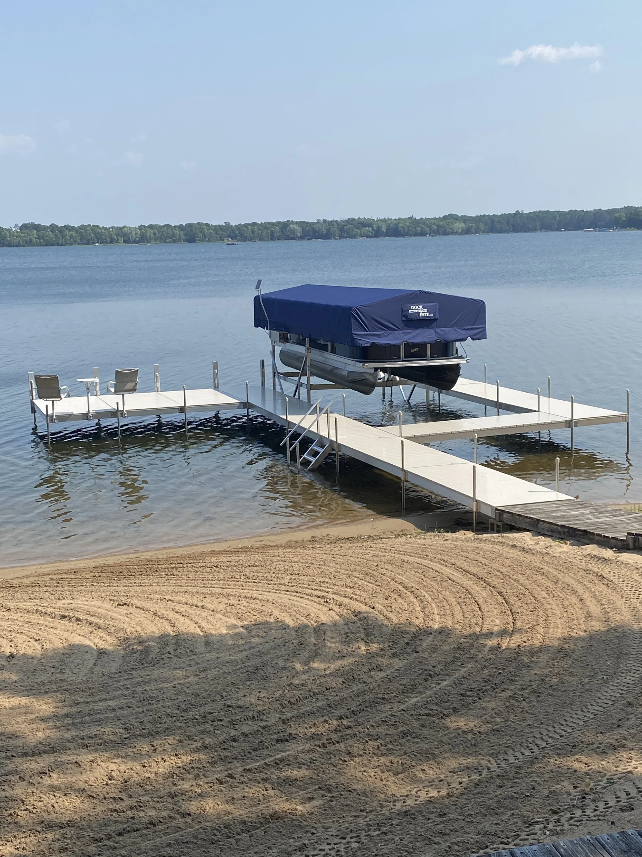 A boat dock with a covered boat, surrounded by water and a sandy shore in the foreground, with a line of trees across the lake in the background under a partly cloudy sky.