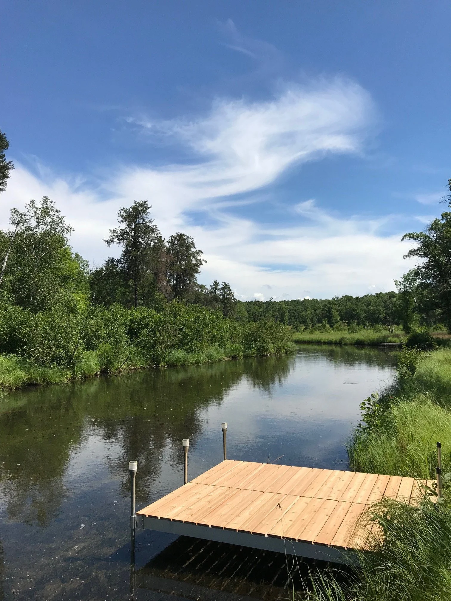 A wooden dock extending into a calm river surrounded by lush green trees, with a blue sky and wispy white clouds overhead.