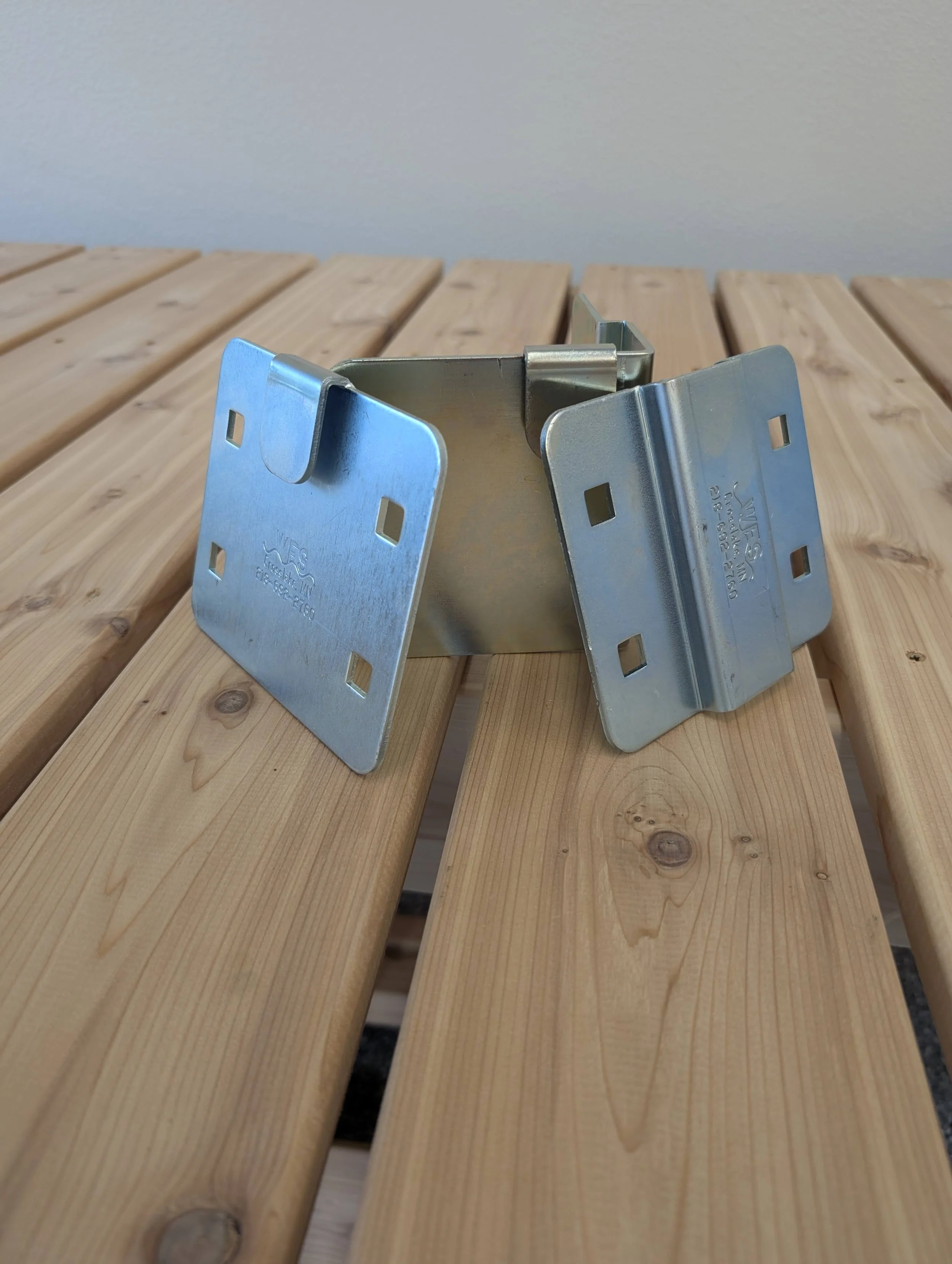 Close-up of a metal bracket placed on a wooden slatted surface, with a plain gray wall background.