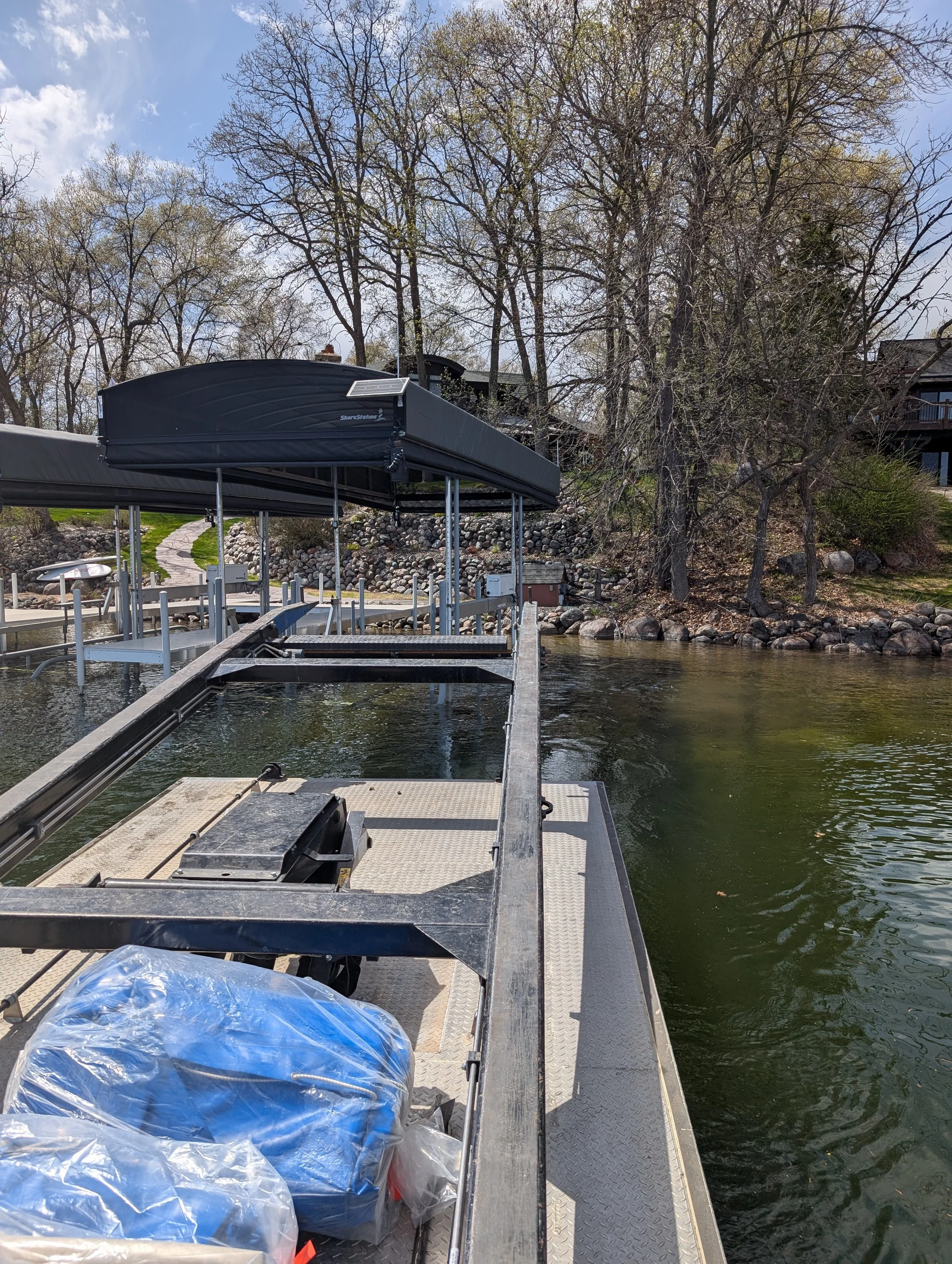 View of a marina dock with a covered boat lift, plastic bags in the foreground, calm water, leafless trees, and houses in the background on a partly cloudy day.