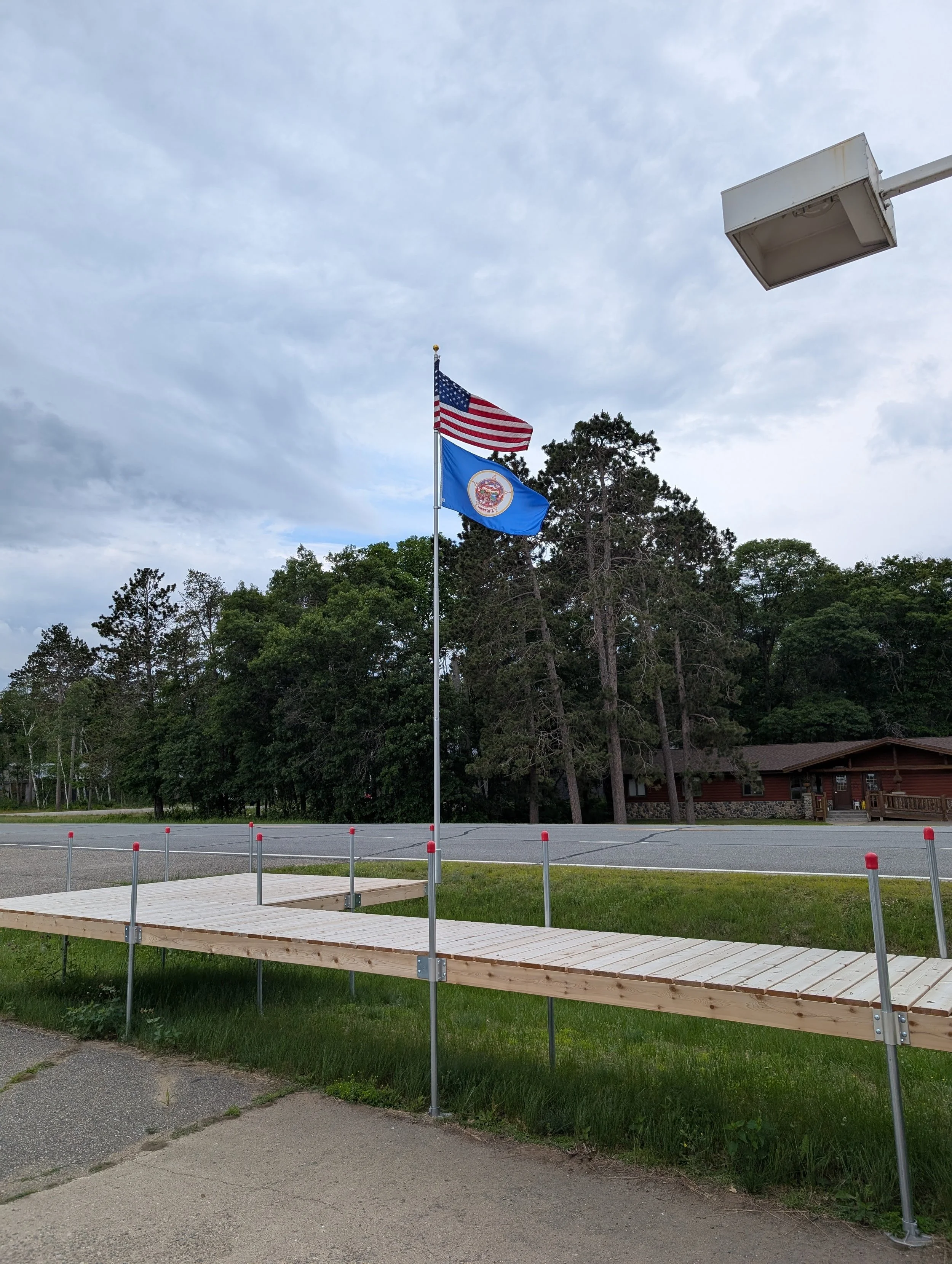 American flag and state flag flying on flagpole in a parking lot with trees and building in the background.