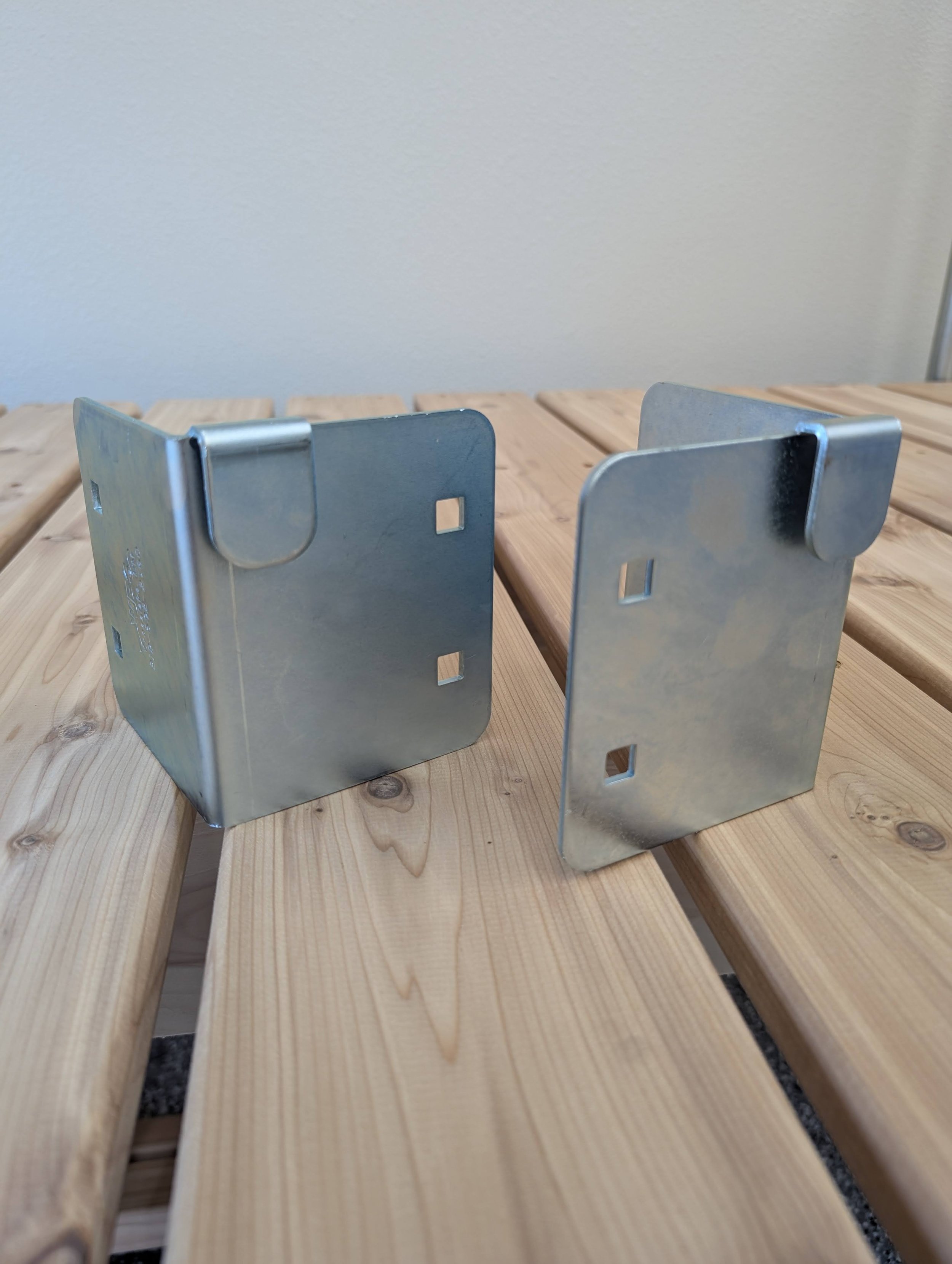 Two metal brackets placed on a wooden slatted surface with a plain wall in the background.