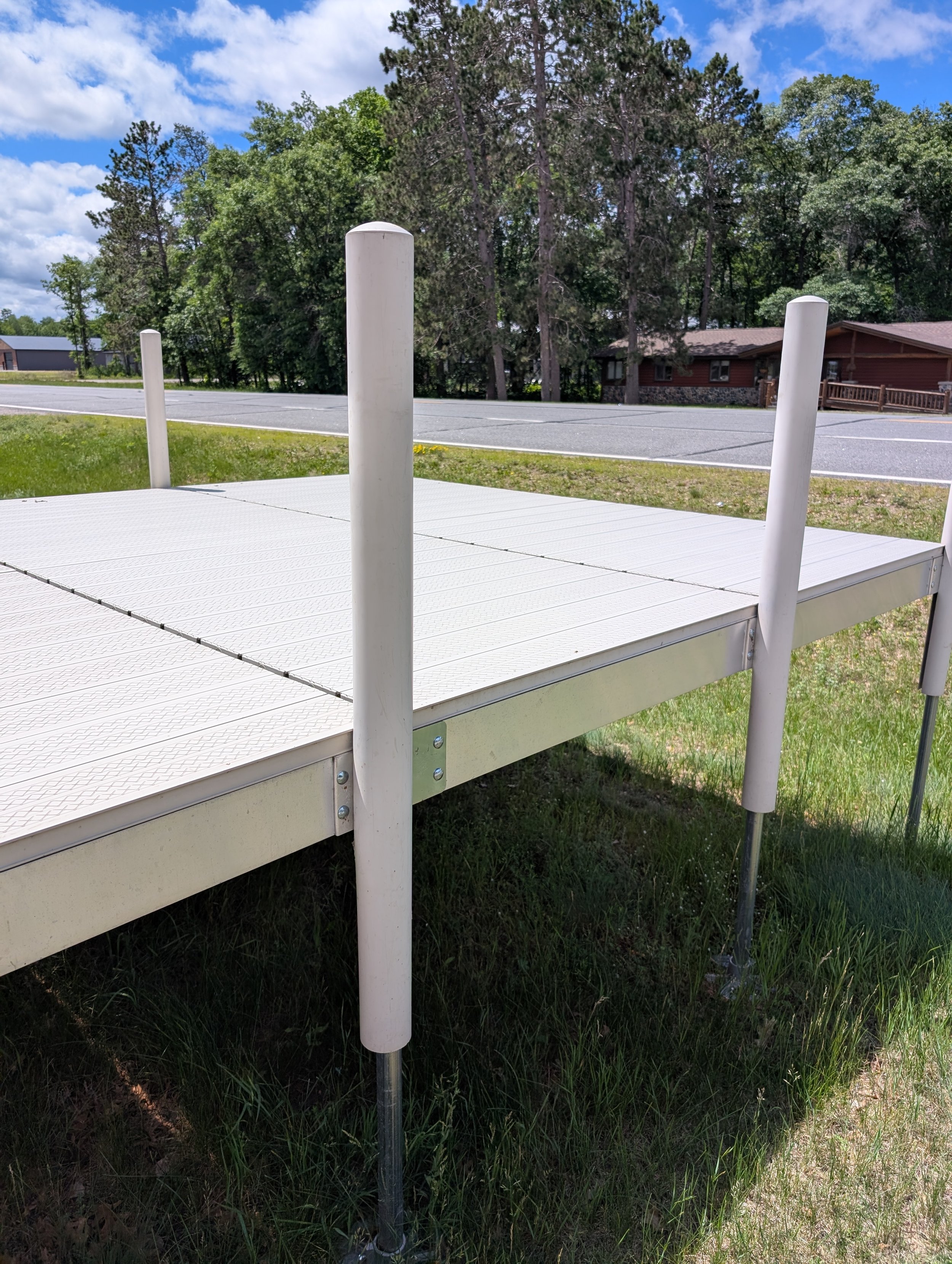 A white platform with three vertical white posts, set on grass near a street. In the background, there are trees, a house, and a partly cloudy sky.