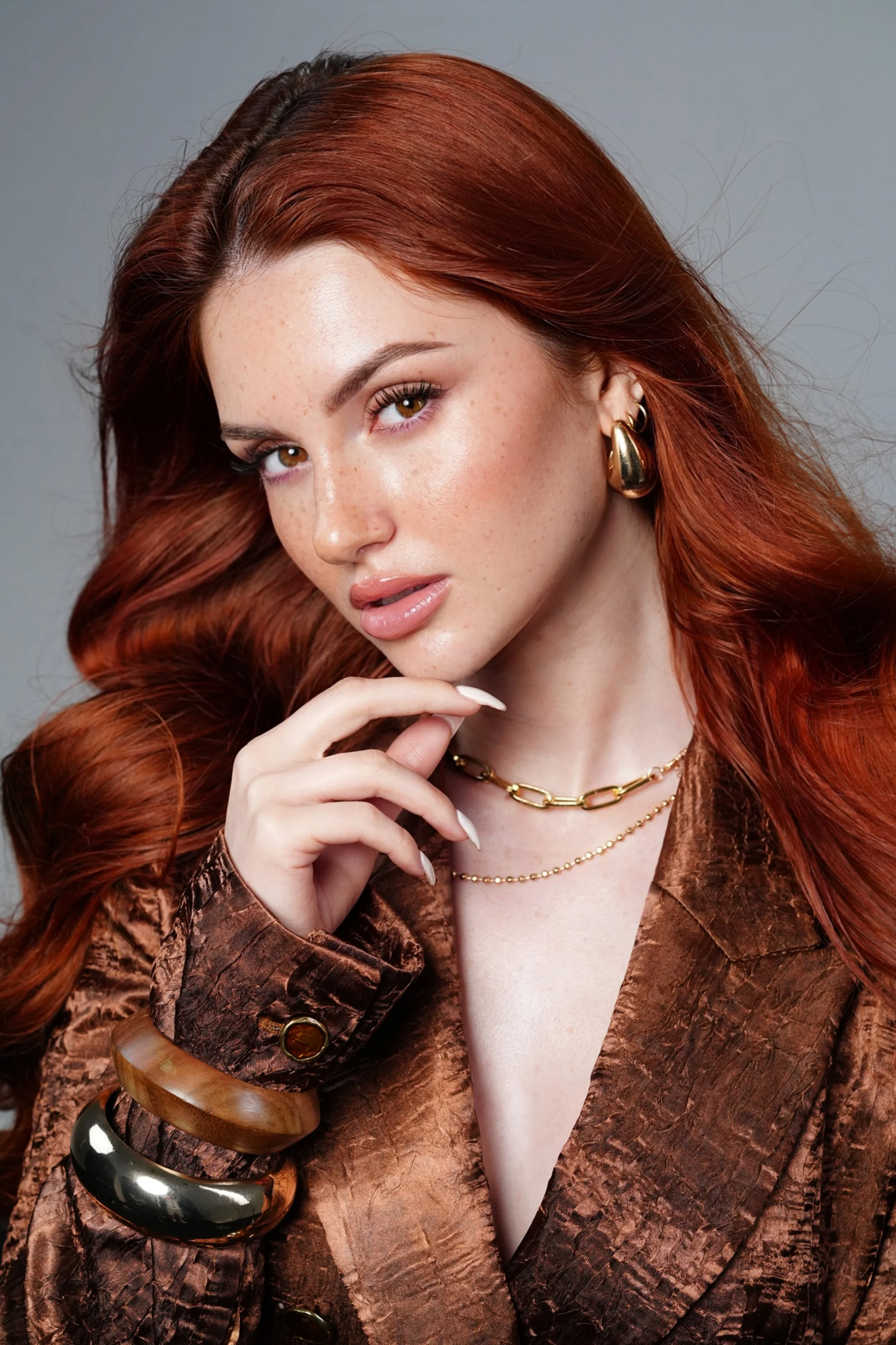 Ragazza con capelli ricci rossi, indossa orecchini dorati, collane dorate, e un abito marrone in tessuto con motivi lucidi, tiene il mento con la mano, sfondo grigio.