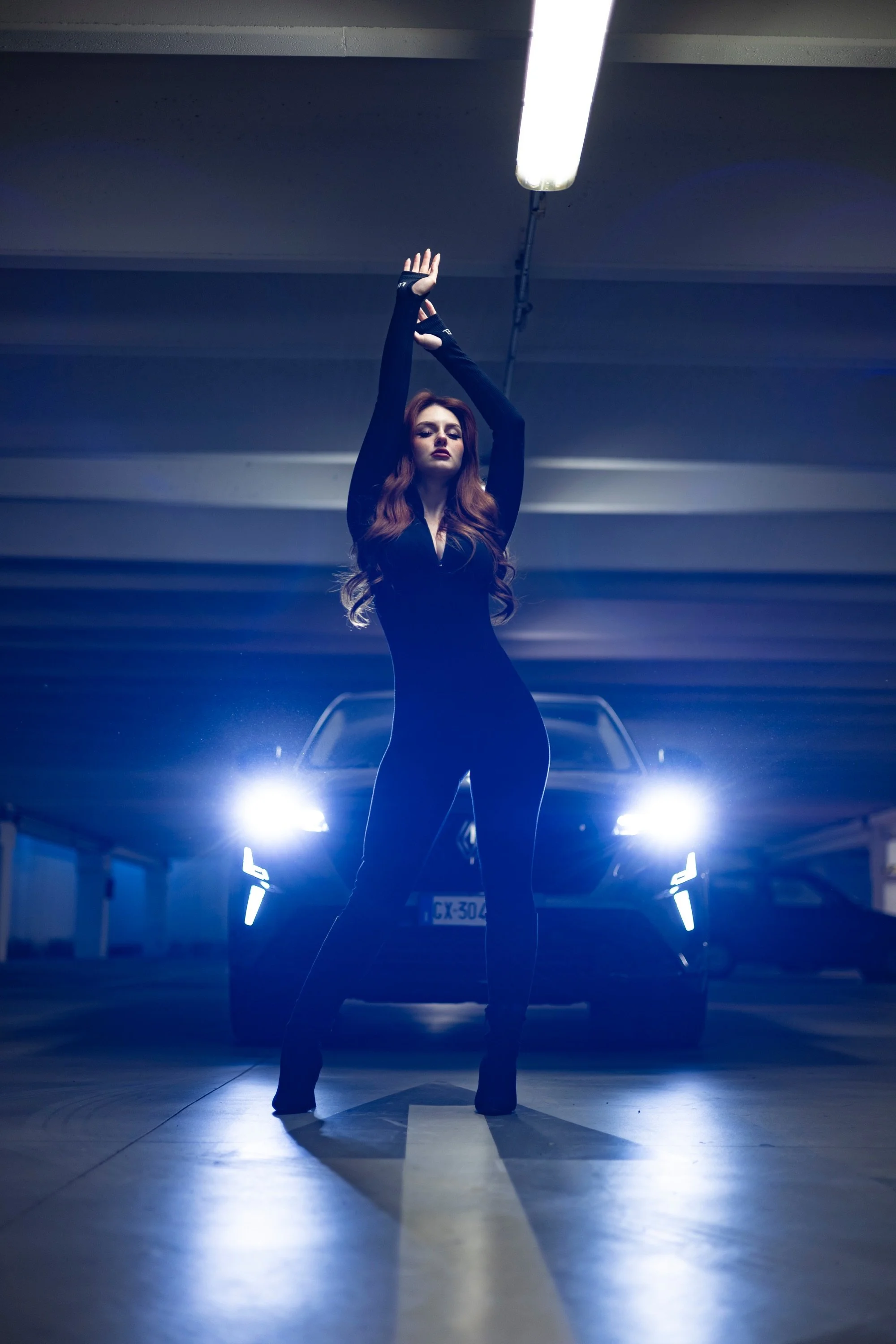 Una donna con lunghi capelli castani scuri e vestito nero posa in un garage con luci al neon. È in piedi davanti a due automobili e la sua postura ha le braccia sollevate sopra la testa.