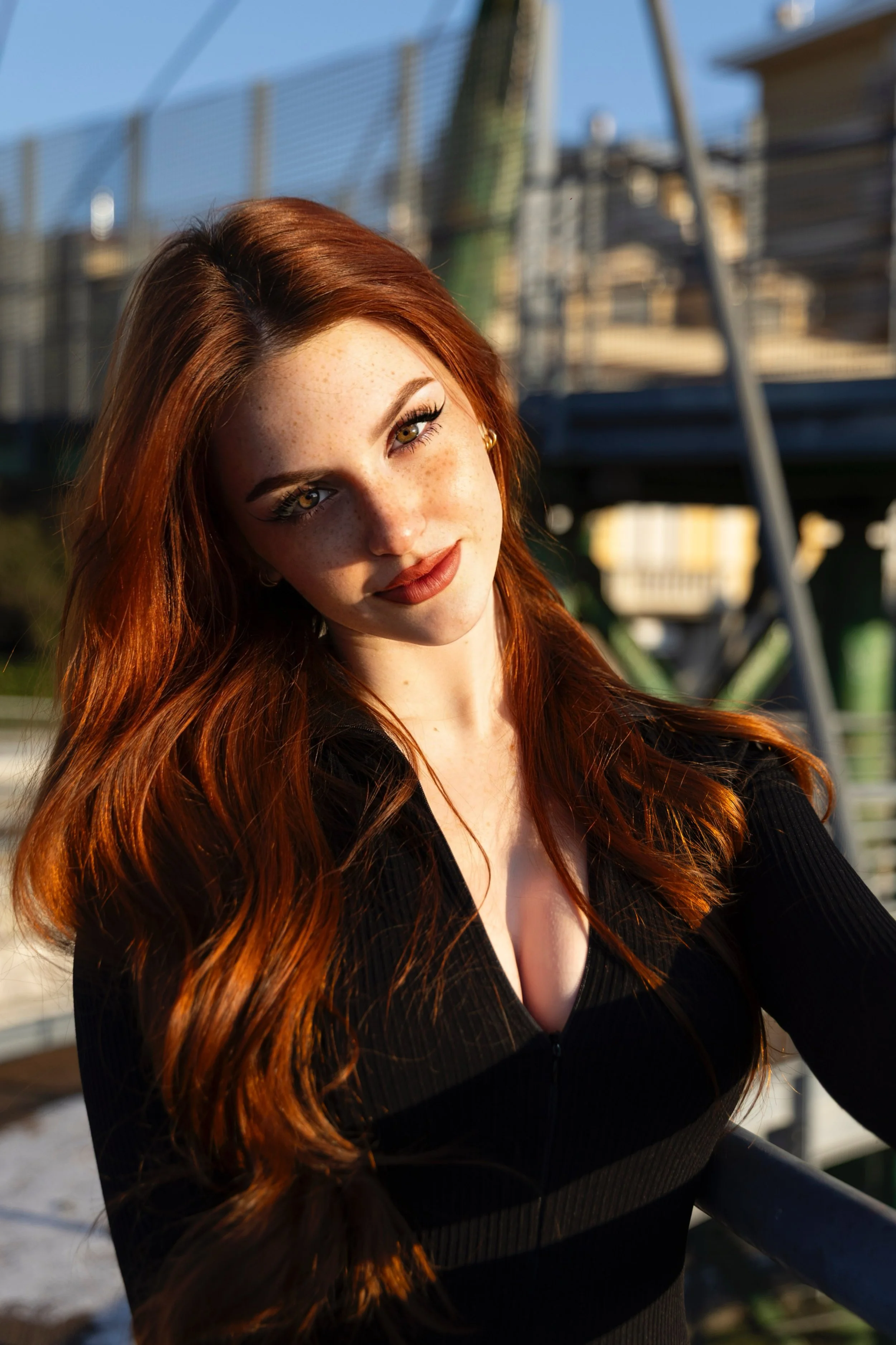 Ragazza con capelli rossi ricci, occhi chiari, che sorride leggermente, indossa un vestito nero a maniche lunghe, in esterna con sfondo di un ponte o ringhiera e edificio, illuminata dal sole.