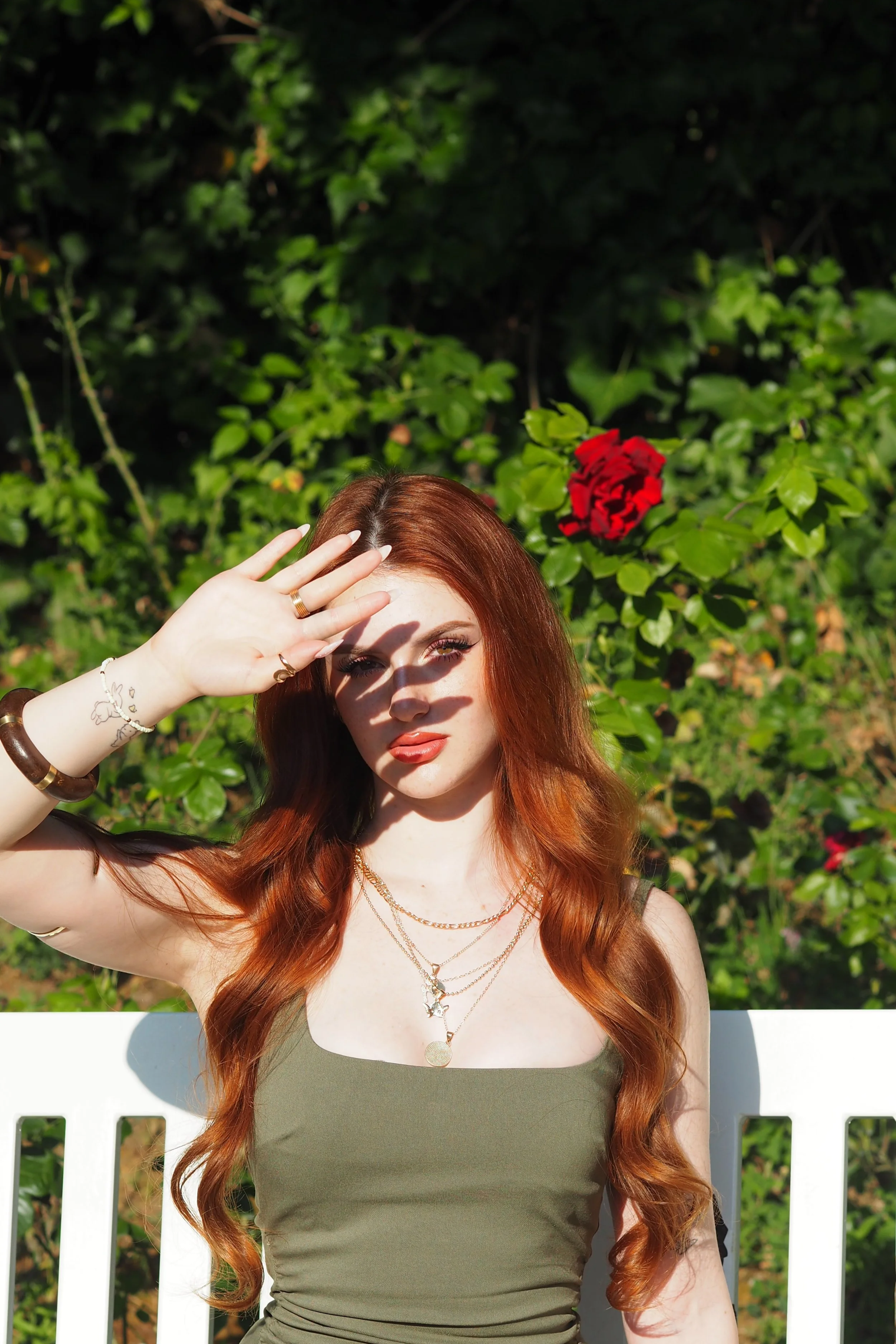 Ragazza con capelli rossi chiari, indossa un top verde oliva, è seduta su una panchina bianca, con un braccialetto e collane d'oro, sventola la mano in ombra sul viso. Sfondo con foglie verdi e rosa rossa.