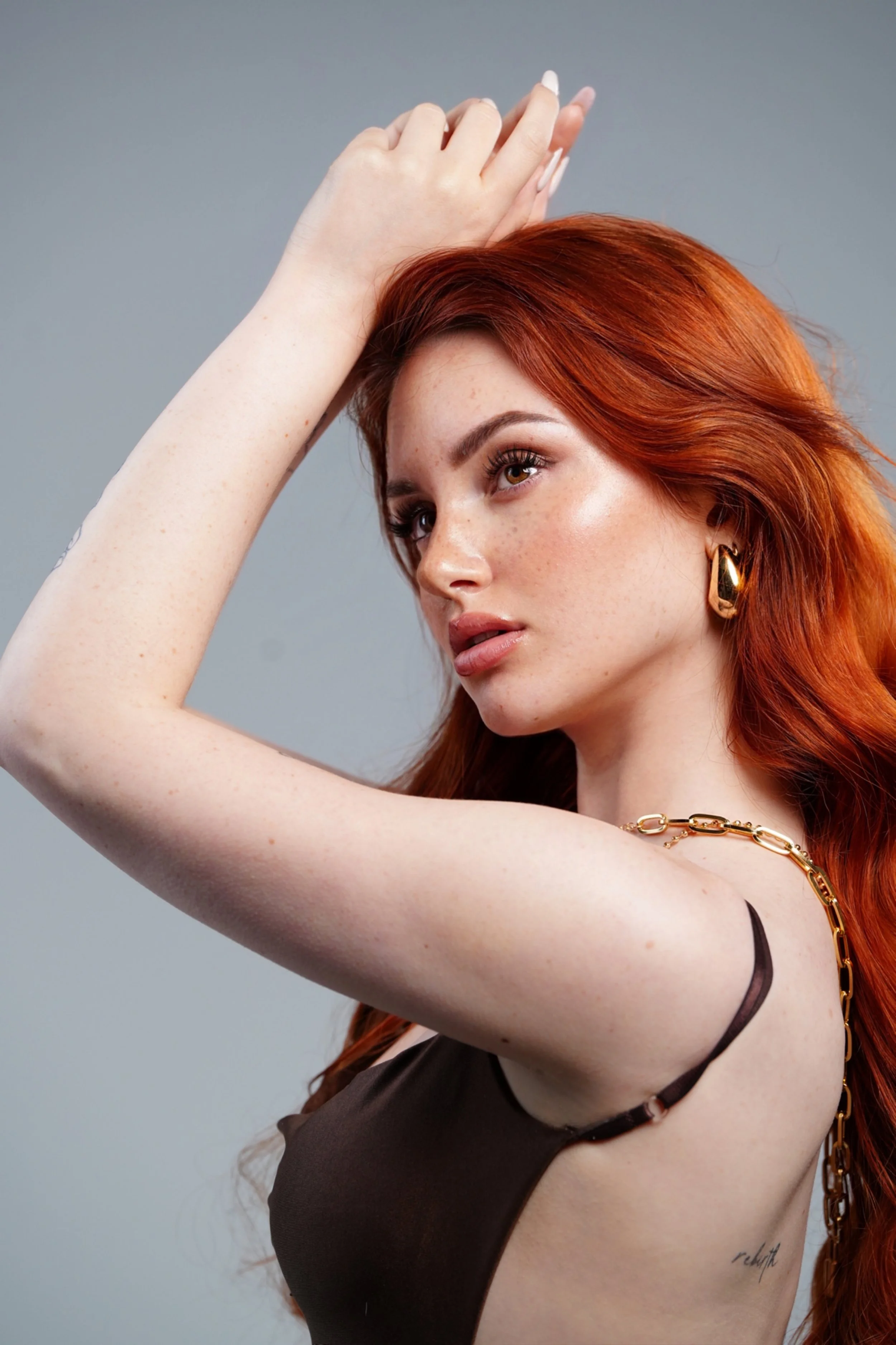 Ragazza con capelli rossi, incinta, indossa orecchini dorati, mostra braccio con tatuaggio sul fianco, vestita con abbigliamento scuro.