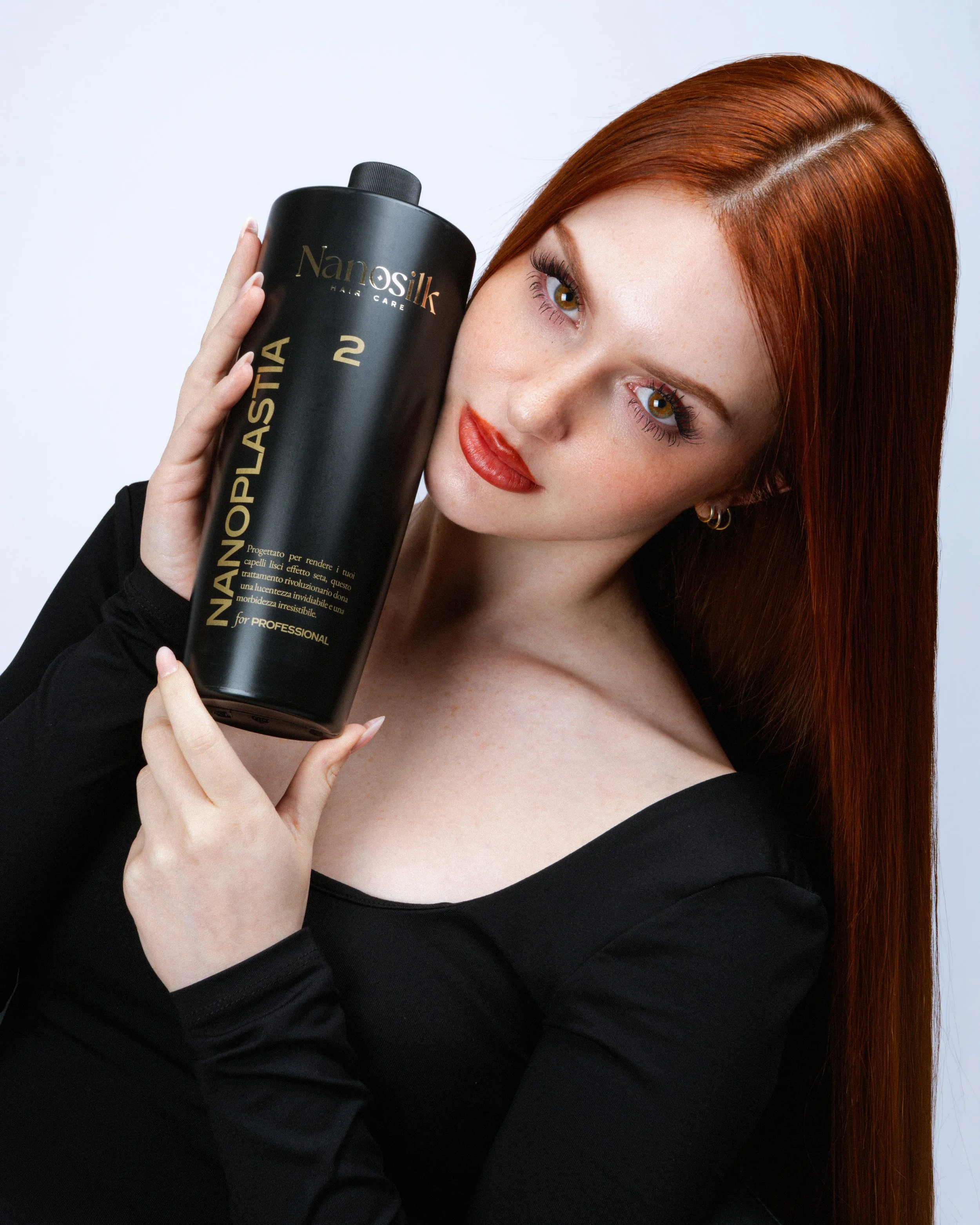 Una donna con capelli rossi, occhi marroni, indossa un abito nero e tiene un contenitore di prodotti per capelli Nanosilk. Lo sguardo è rivolto leggermente di lato, con labbra rosse e trucco leggero.