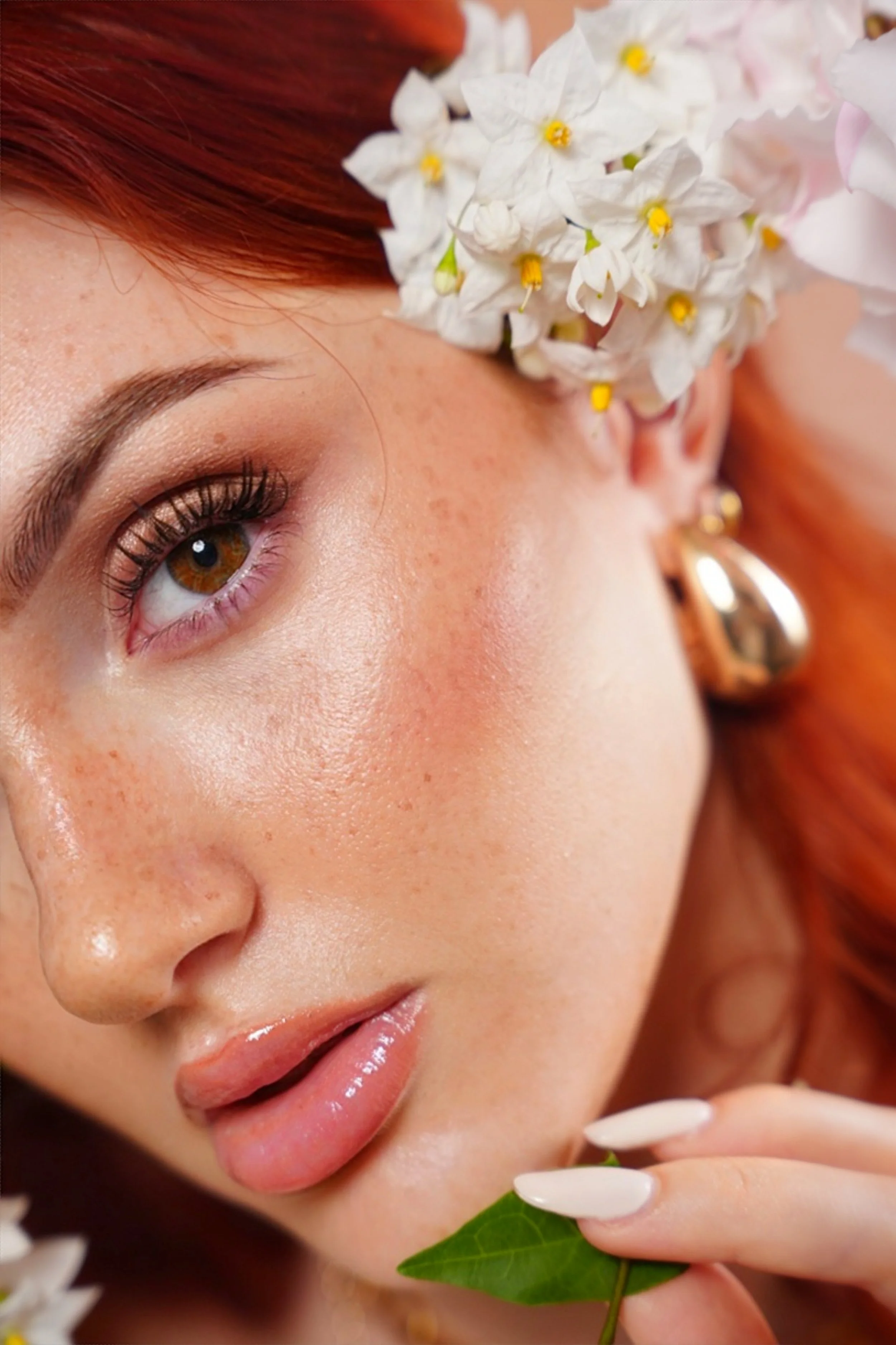 Il primo piano del volto di una donna con la pelle liscia, le labbra leggermente socchiuse, occhi marroni e capelli rossi, con fiori bianchi come decorazione sulla testa, e una foglia verde tra le dita.