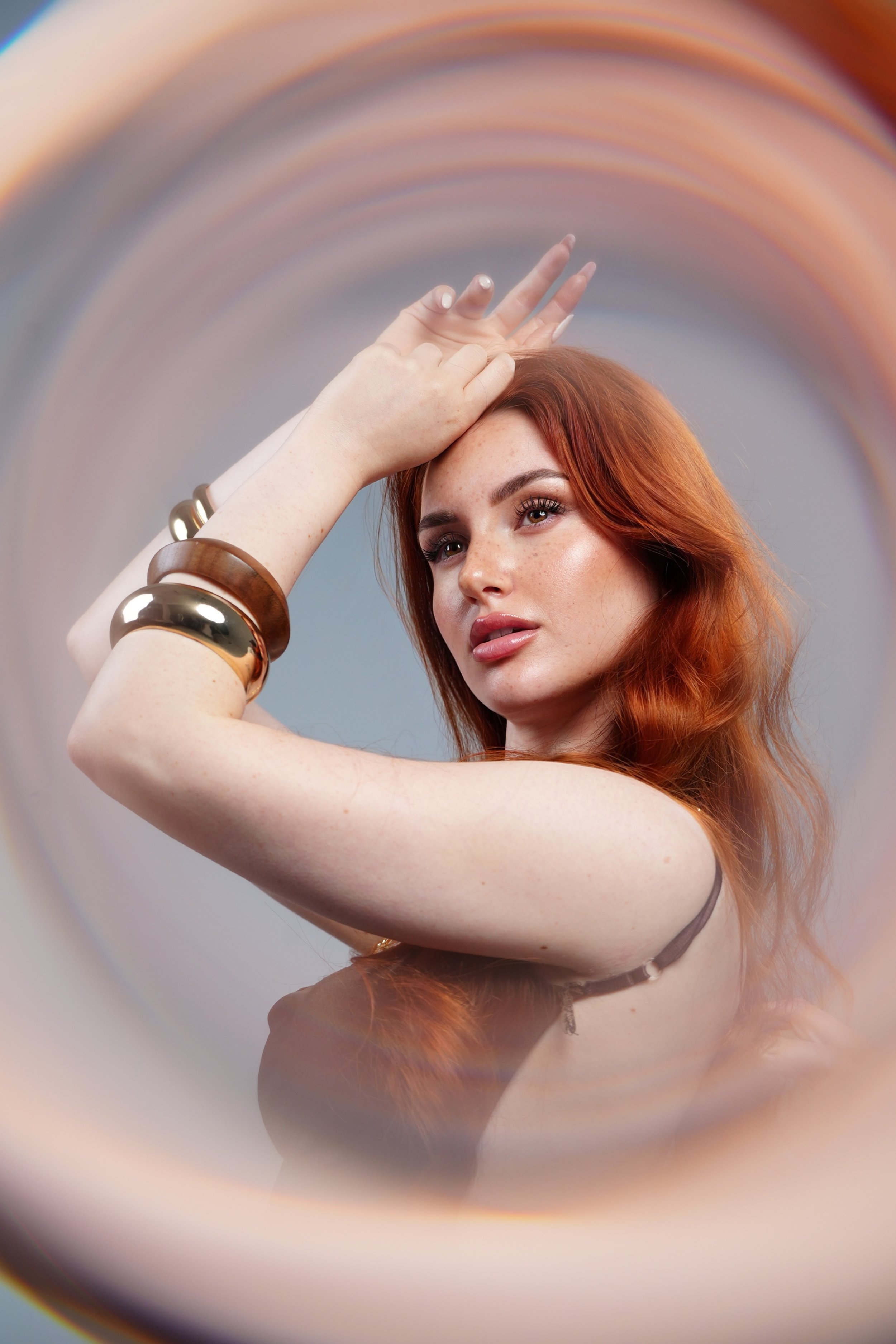 Ragazza dai capelli ricci e rossi, con pelle chiara e lentiggini, che si tocca i capelli con entrambe le mani, indossa bracciali metallici e una maglietta nera, davanti a uno sfondo sfocato con un effetto a tunnel.