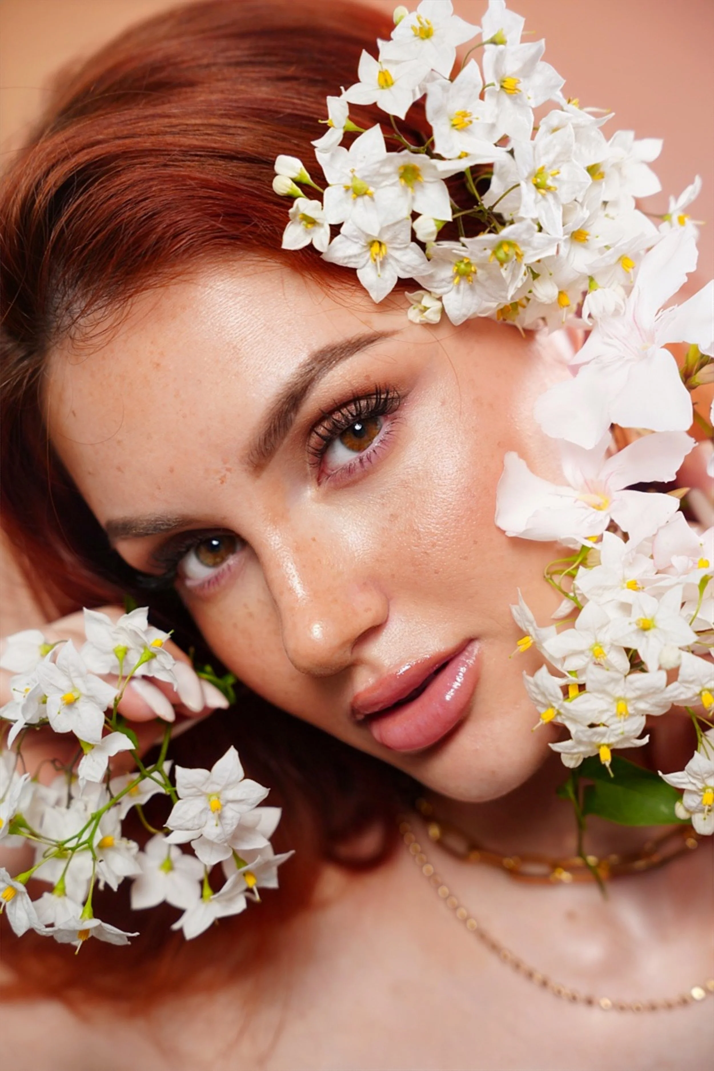 Un primo piano di una donna con capelli castano rame, contornata da fiori bianchi. La donna ha la pelle chiara, occhi castani e labbra carnose, con un leggero sorriso. Indossa una collana dorata e ha un trucco naturale.