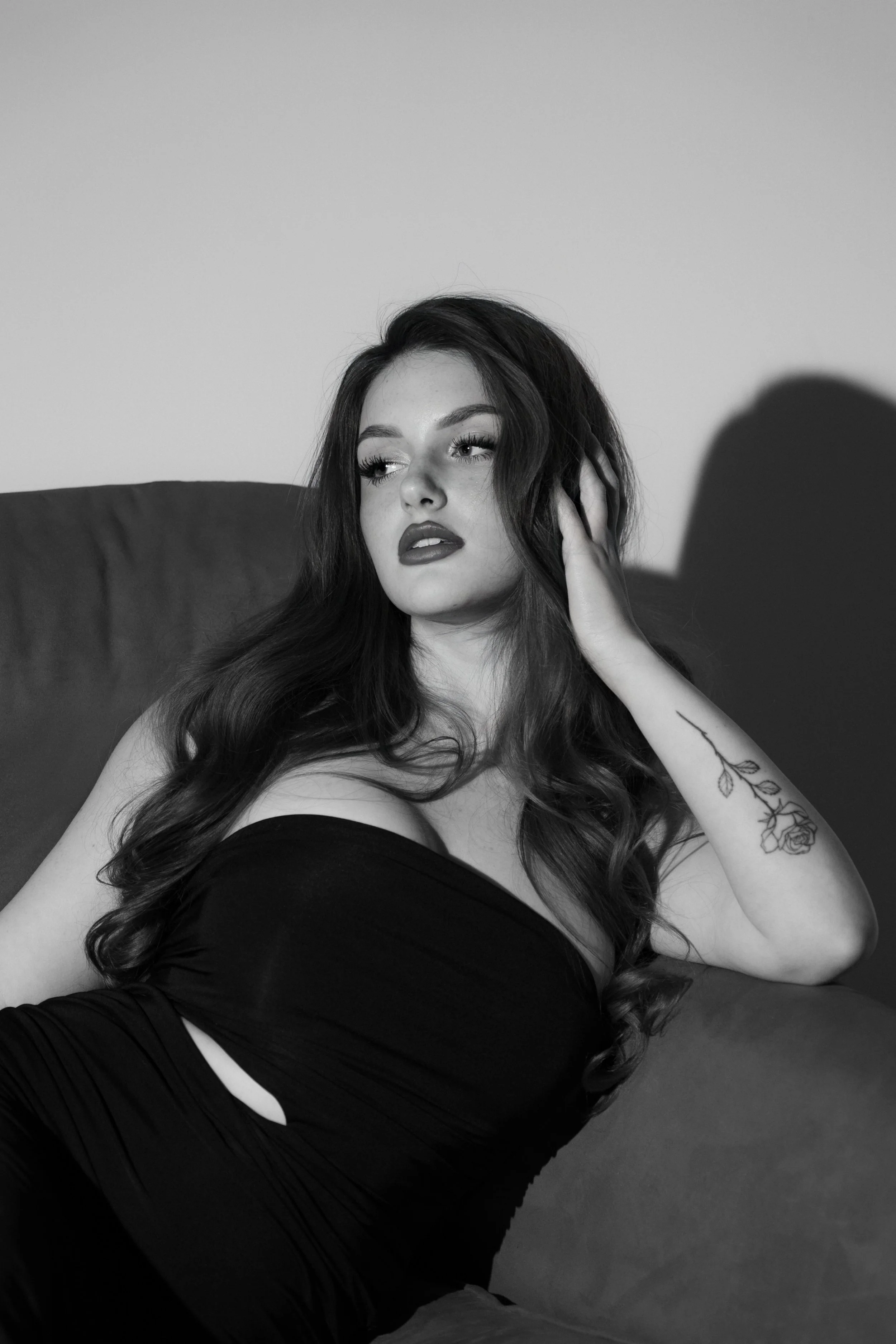 Ragazza con capelli lunghi e ricci, tatuaggio di fiori sul braccio sinistro, indossa un abito nero e si passa una mano sulla testa, seduta su un divano.