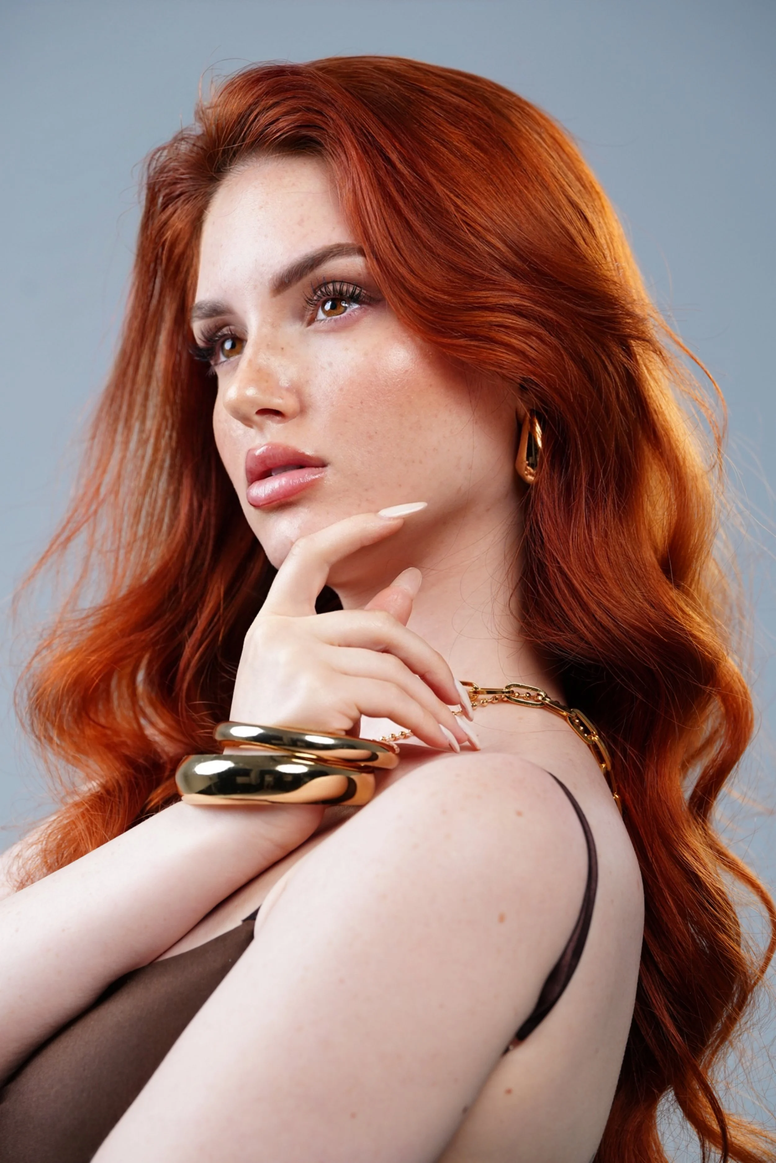 Ragazza con lunghi capelli ricci di colore rosso, abbigliamento in toni neutri, con orecchini d'oro e bracciali dorati. Fa una smorfia pensierosa e appoggia il mento sul dito indice mentre guarda lontano. Sfondo grigio chiaro.