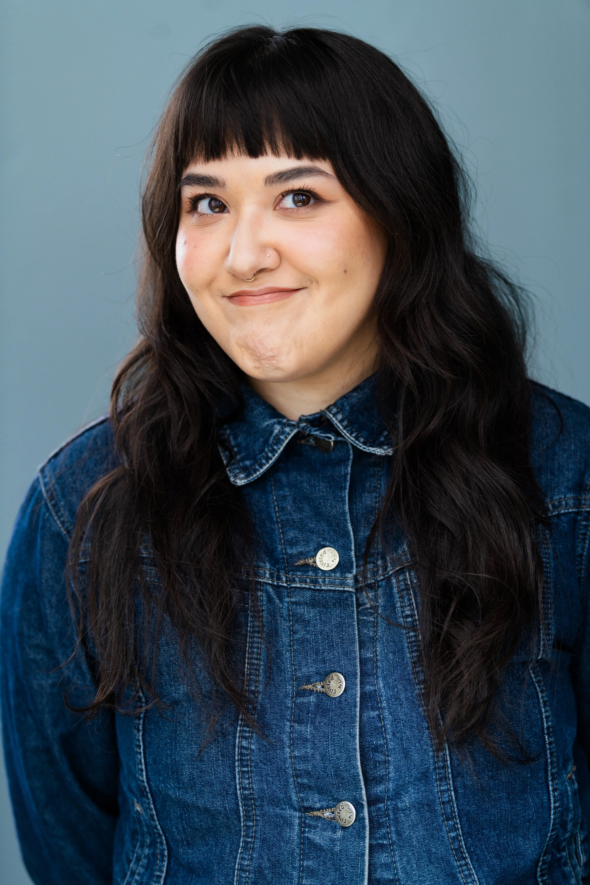 A woman with long dark wavy hair, wearing a denim jacket with metal buttons, smiling and looking at the camera against a plain background.