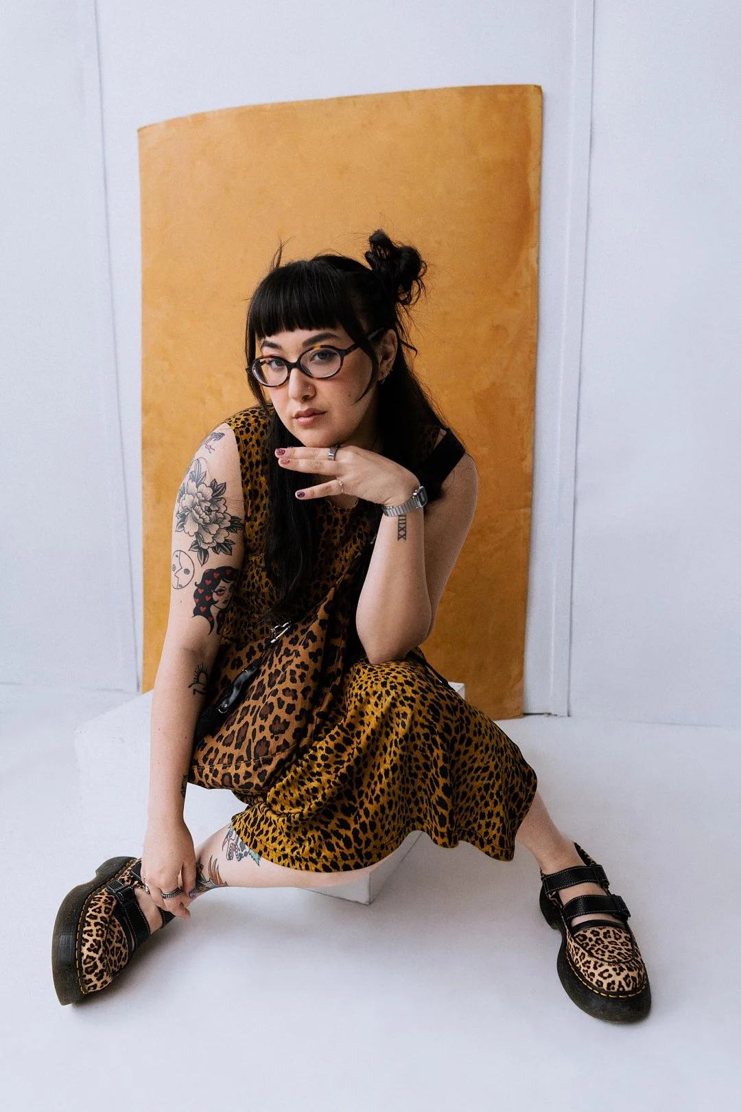 Young woman with black hair and glasses sitting on a white platform, wearing a leopard print dress and shoes, with tattoos on her arm and leg, posing against a wooden backdrop.