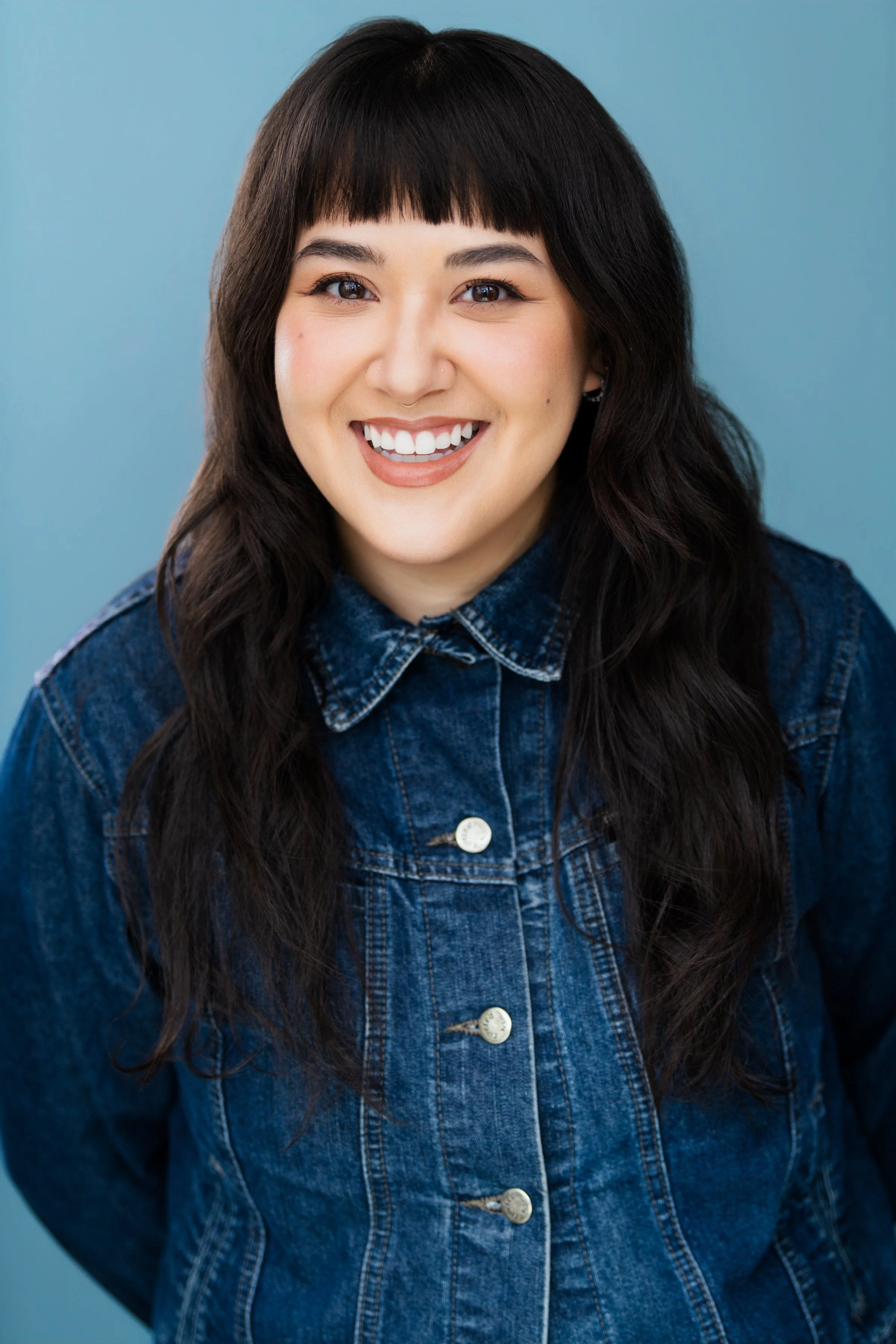 Close-up of a woman with long dark hair, wearing a denim jacket, smiling against a light blue background.