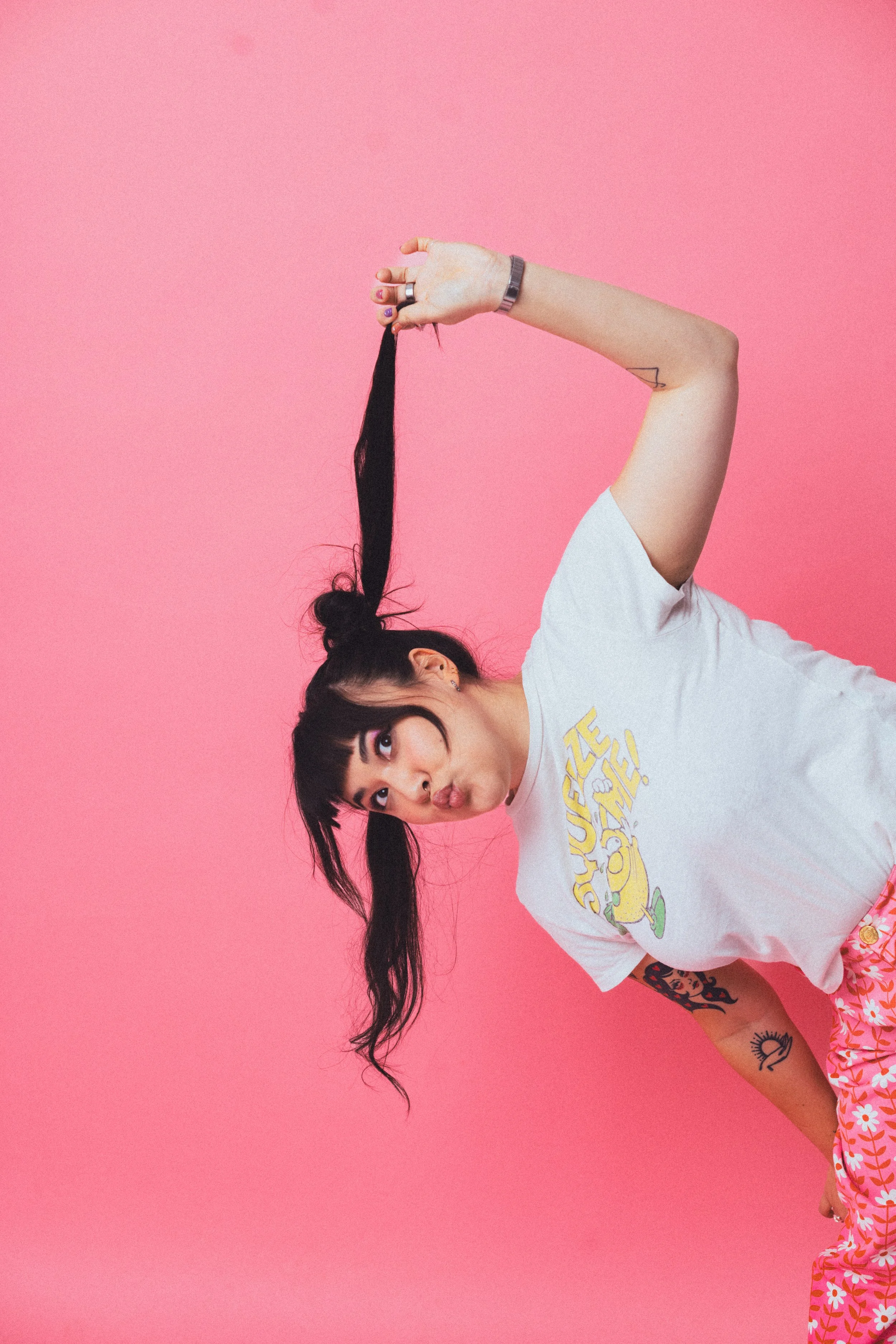 Young woman with long black hair making a funny face, sticking out her tongue, against a pink background. She is wearing a white t-shirt with a cartoon character and pink floral pants.