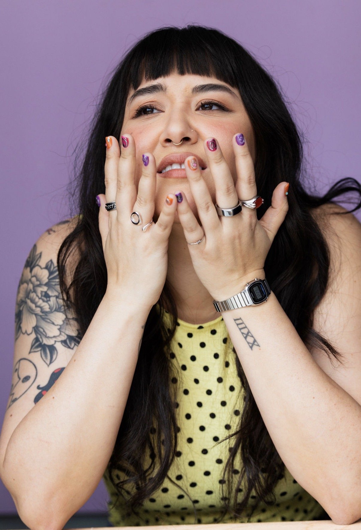 A young woman with dark hair, tattoos on her arms, and multiple rings on her fingers, touching her face with a contemplative expression, wearing a yellow polka-dot top and a silver watch, against a purple background.