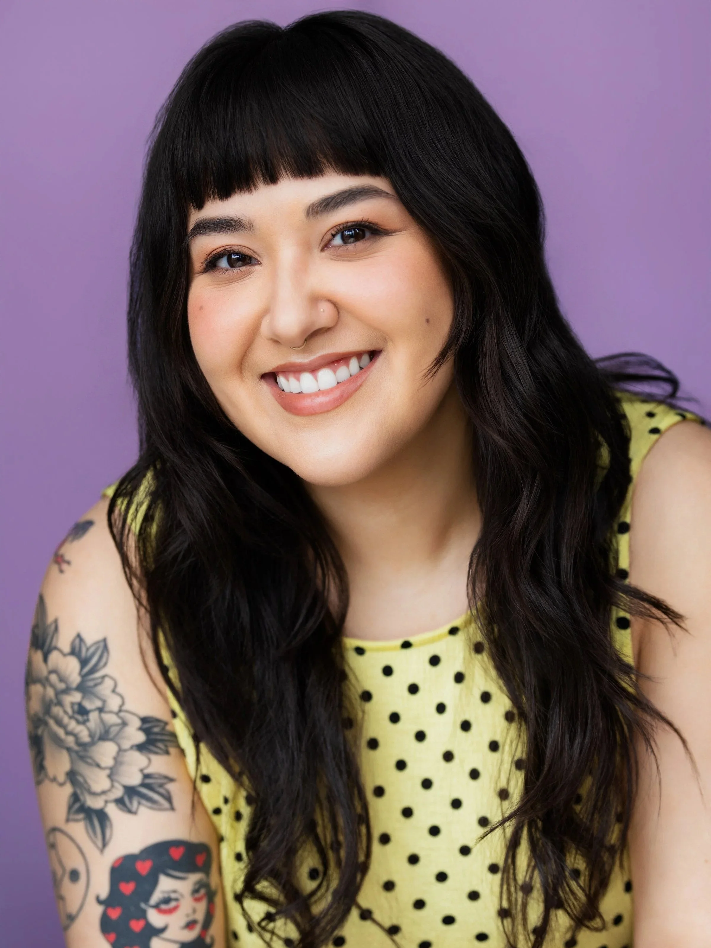 A young woman with long dark hair and blunt bangs smiling in front of a purple background, wearing a yellow sleeveless top with black polka dots, and has tattoos on her left arm including a woman's face with hearts and floral designs.