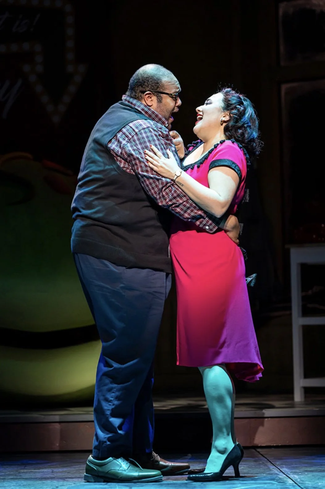 A man and woman share a joyful moment on stage, smiling and holding each other. The woman is wearing a pink dress with black accents, high heels, and has short hair. The man is dressed casually in a plaid shirt, vest, and slacks. The background is da
