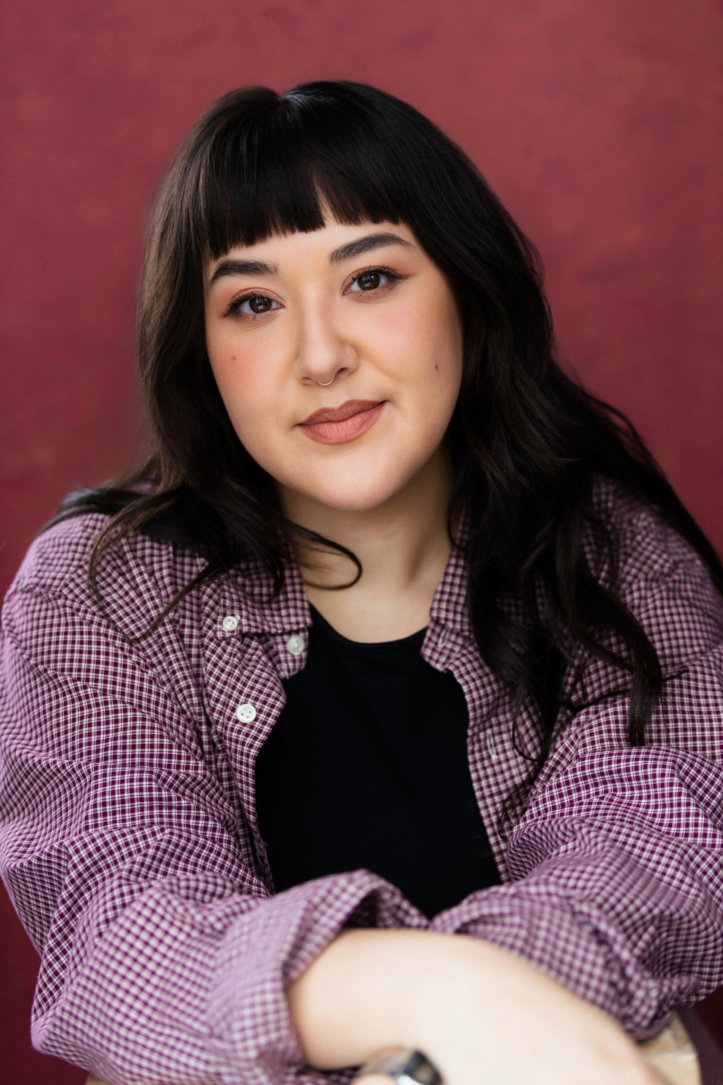 A young woman with black hair and a septum piercing, wearing a checkered shirt over a black top, sitting with her arms crossed against a pinkish maroon background.