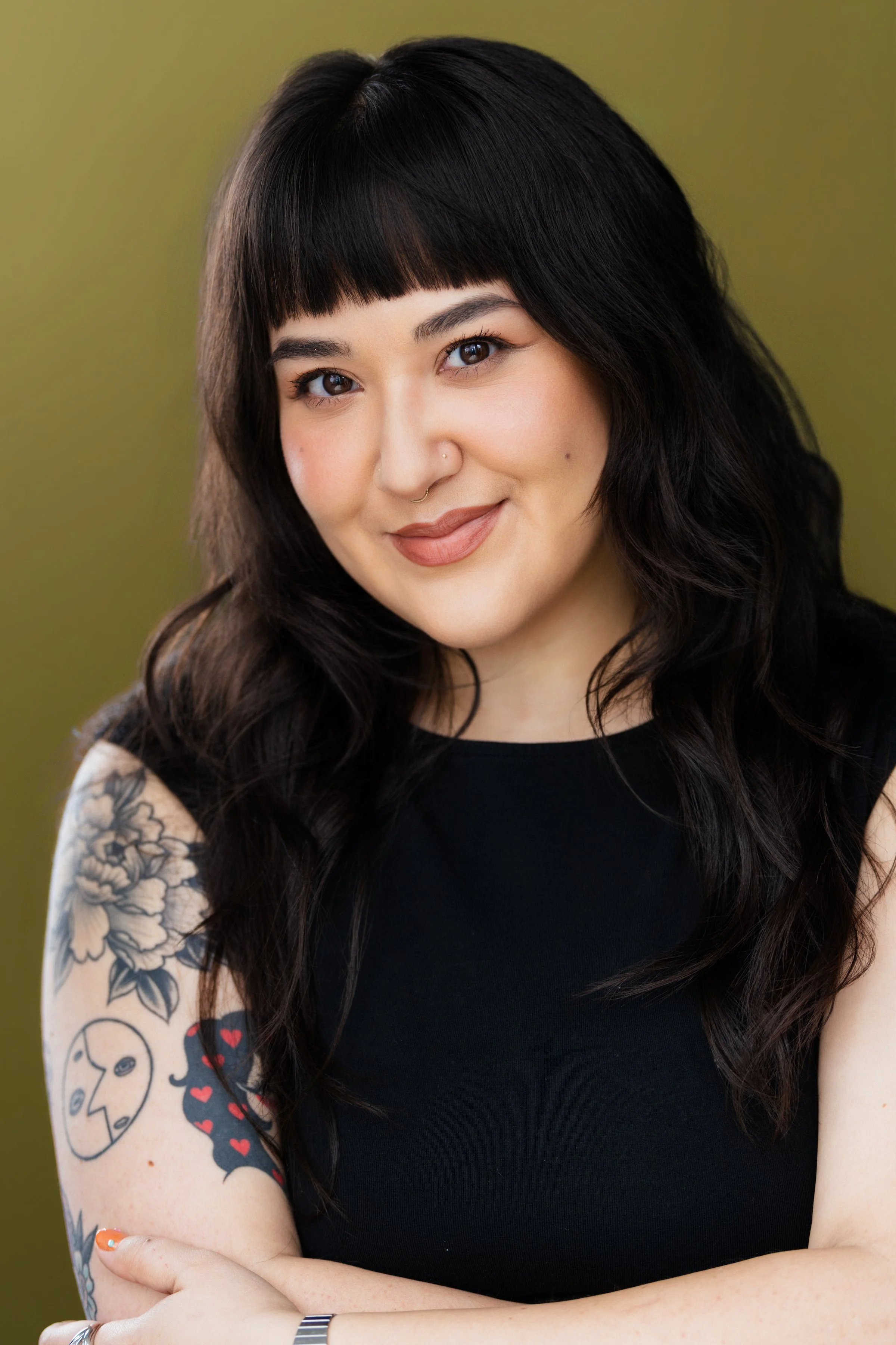 A woman with long dark wavy hair and bangs, wearing a black top, with tattoos on her arm, smiling at the camera against a green background.